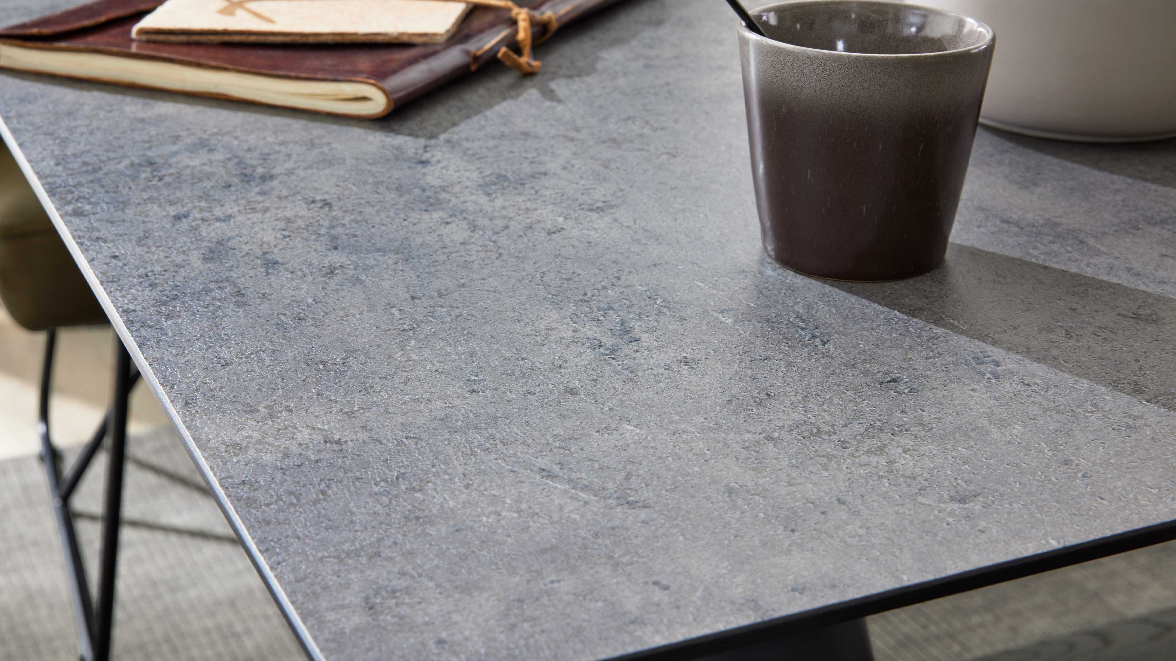 Grau strukturierte Tischplatte in Steinoptik mit brauner Keramiktasse rechts, Ledernotizbuch oben links und schwarzem Metallgestell.