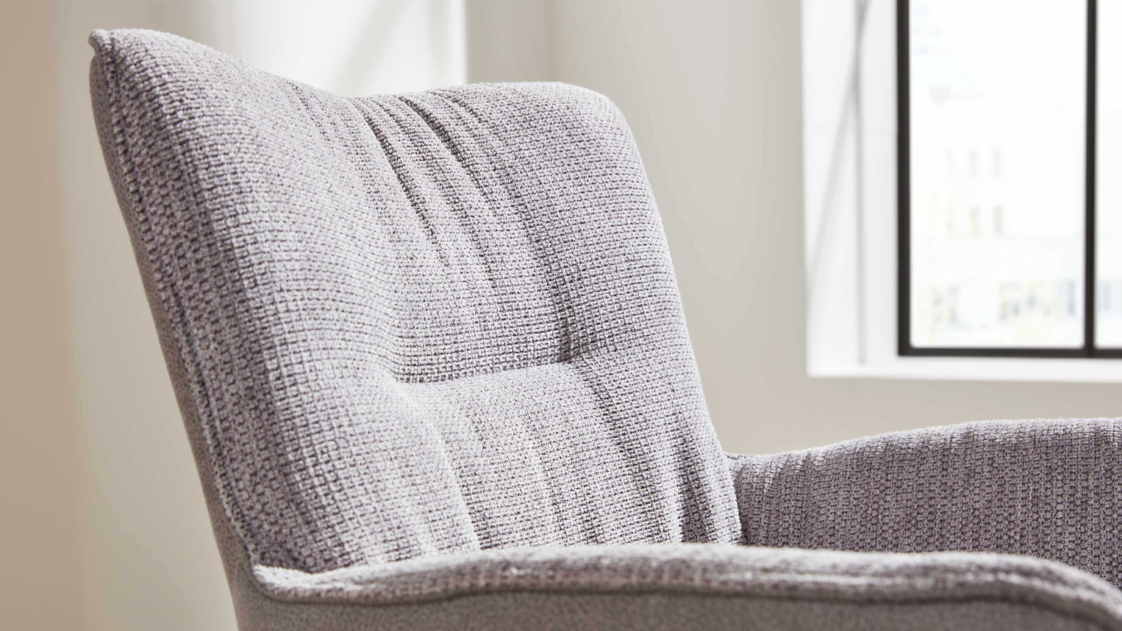 Polsterlehne und Arm eines hellgrauen Sessels mit grob gewebter Struktur und sichtbaren Steppnähten, Fenster dahinter.