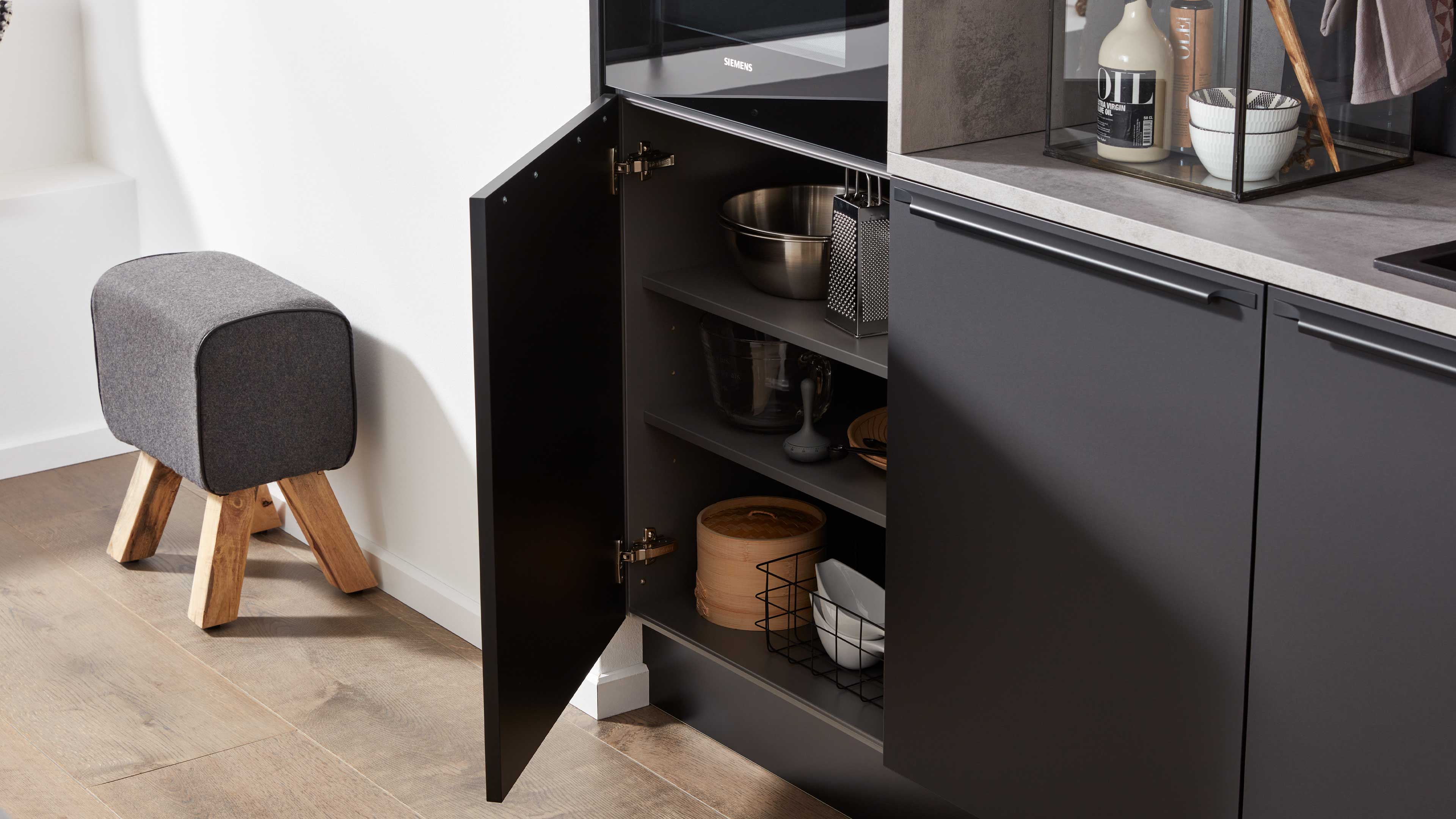 Offener dunkelgrauer Küchenschrank mit Schüssel und Reibe, Glas- und Holzschalen sowie Drahtkorb; links gepolsterter Hocker mit Holzbeinen.
