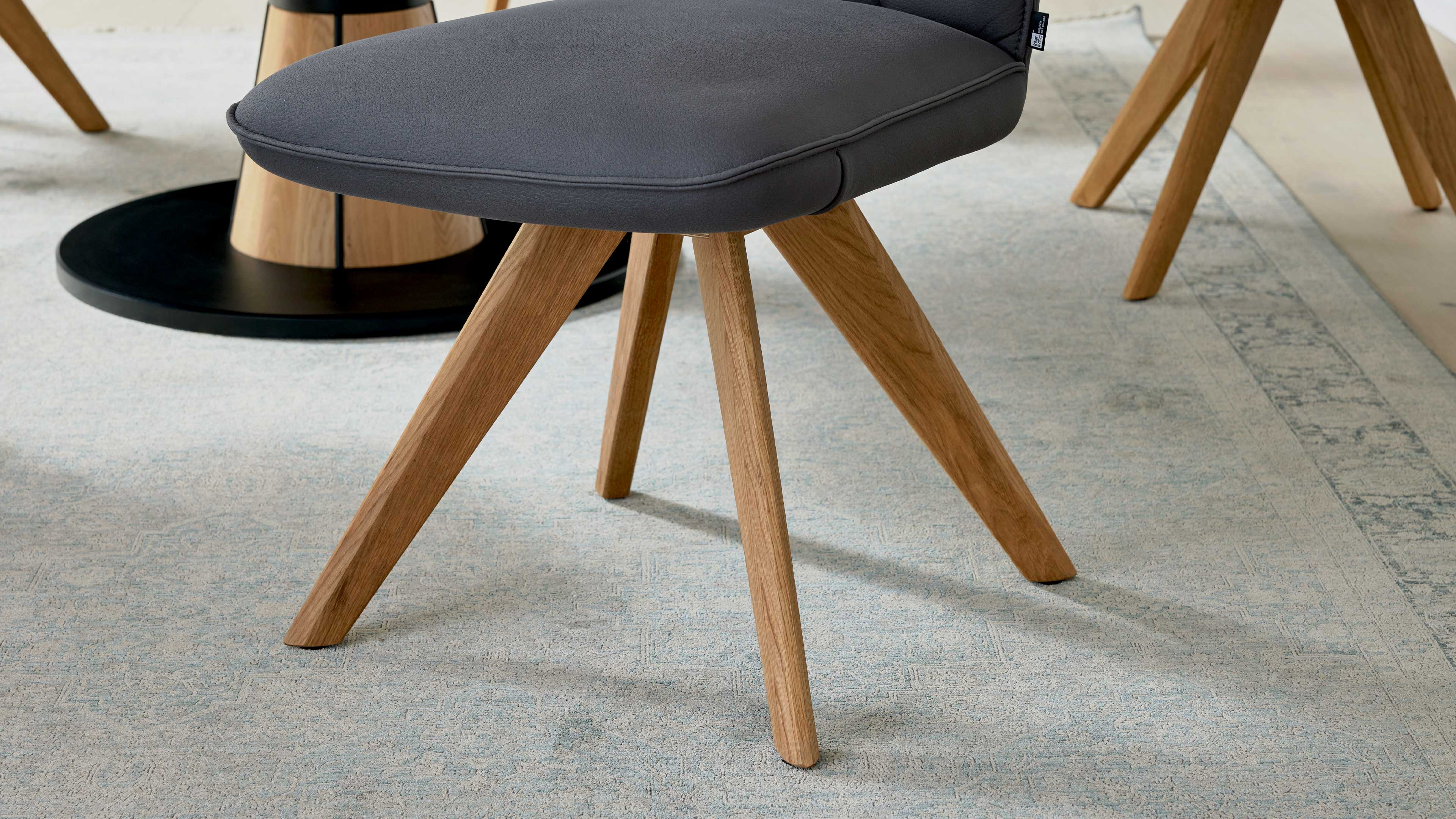 Grauer, gepolsterter Stuhlsitz auf vier nach außen geneigten, holzfarbenen Beinen, auf hellem gemustertem Teppich neben schwarzem Tischfuß.