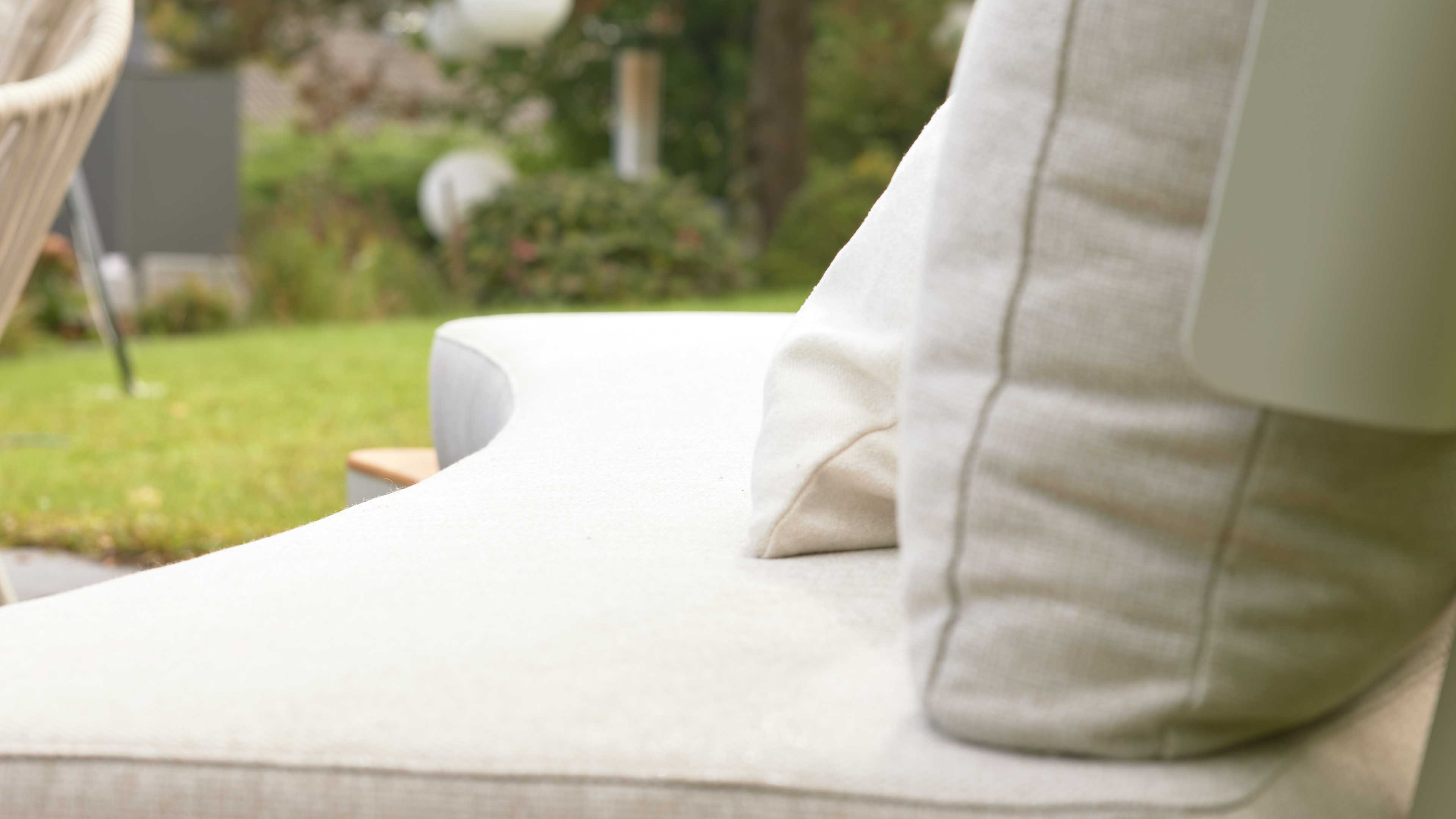Helles Detail einer gebogenen Sofa-Sitzfläche mit zwei beigefarbenen Rückenkissen und unscharfem Gartenhintergrund.