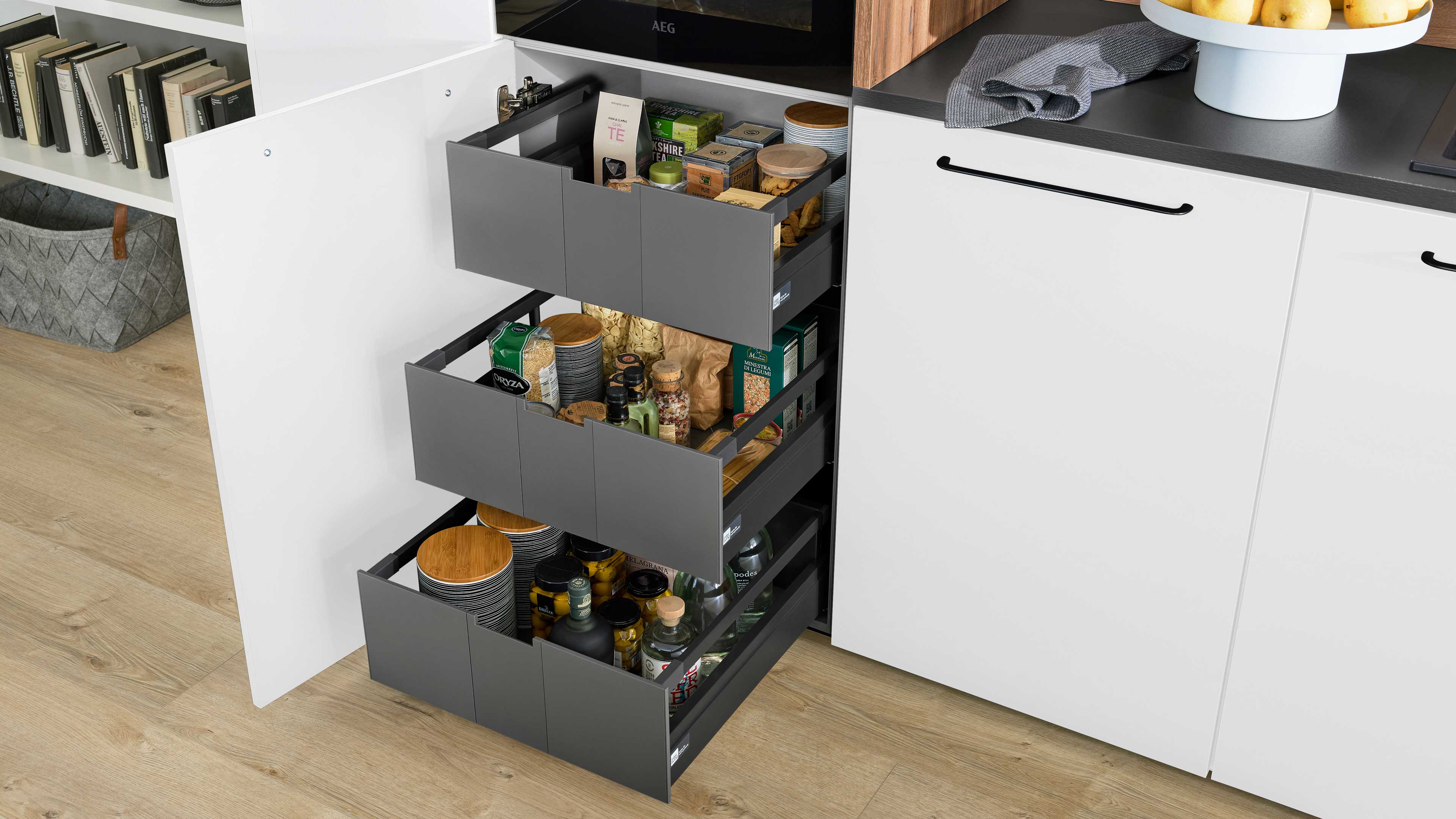 Drei graue Auszugsboxen in offenem Küchenunterschrank mit eingeordneten Vorratsdosen, Flaschen und Gläsern.
