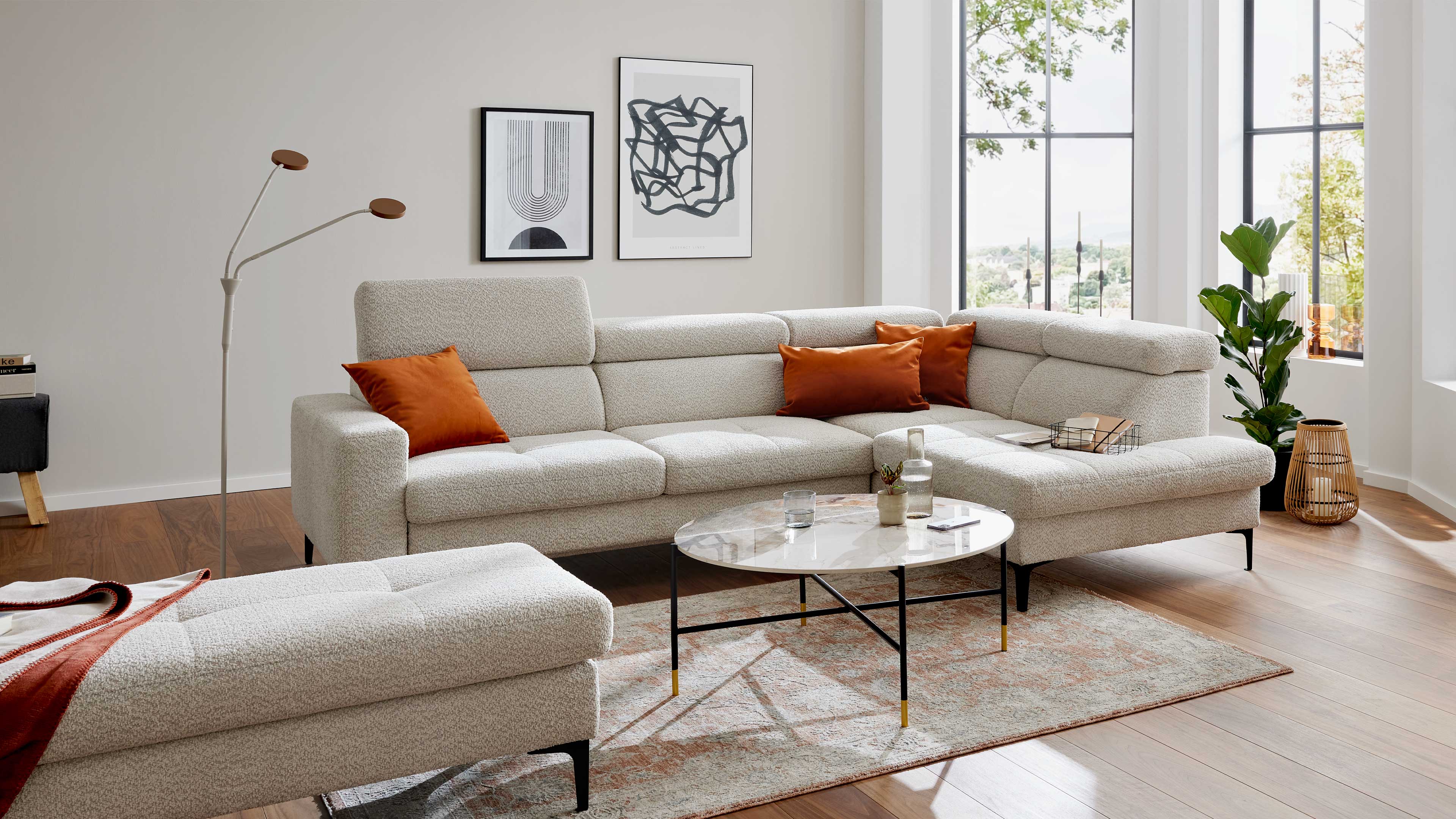Hellbeiges Ecksofa mit orangefarbenen Kissen, runder Marmor-Couchtisch, Fußbank, Stehleuchte, Bilder an der Wand und Pflanzen.