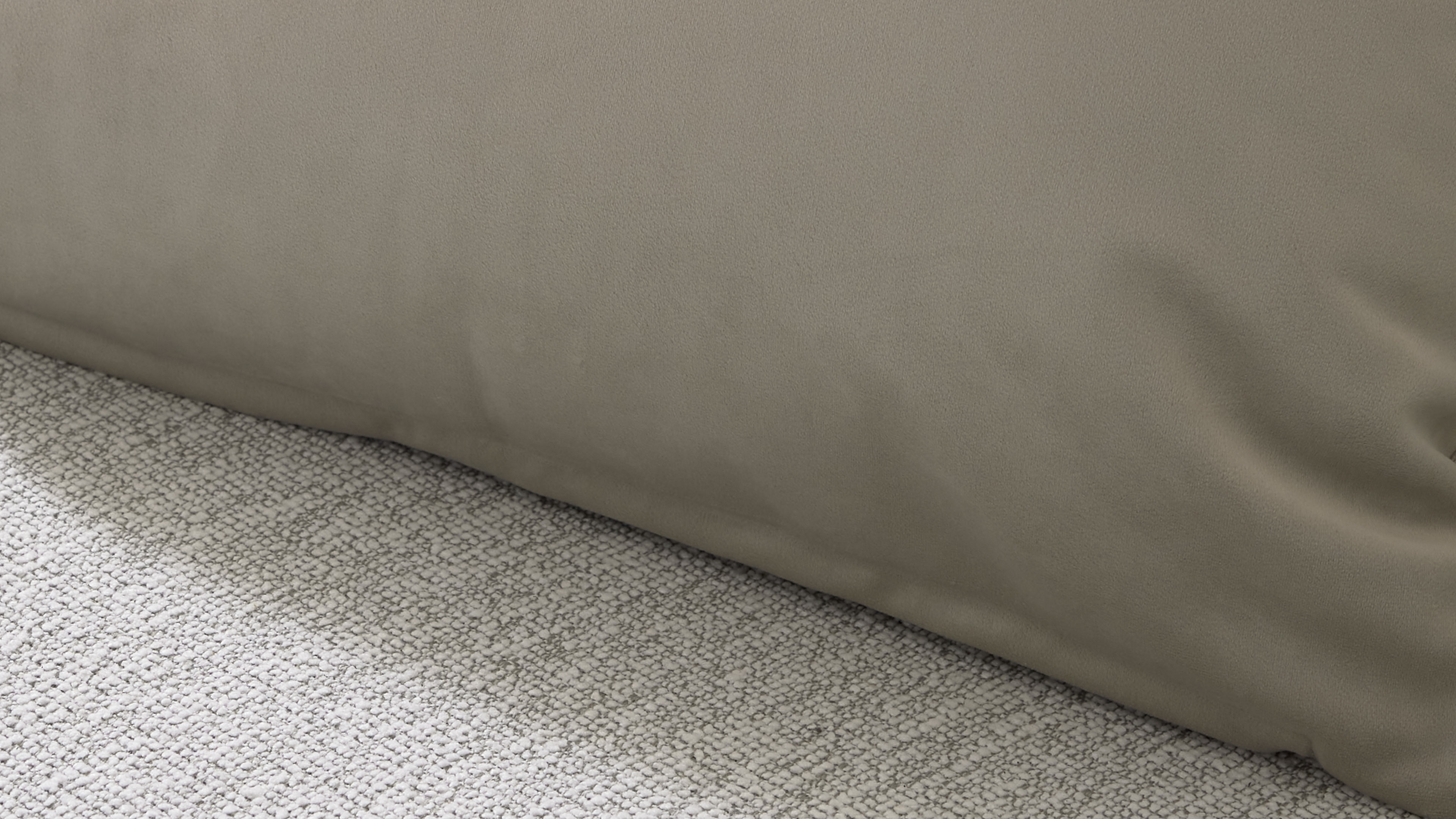 Unterkante eines taupefarbenen Stoffbezugs, der leicht Falten wirft, über einem hellgrauen, strukturierten Teppich.