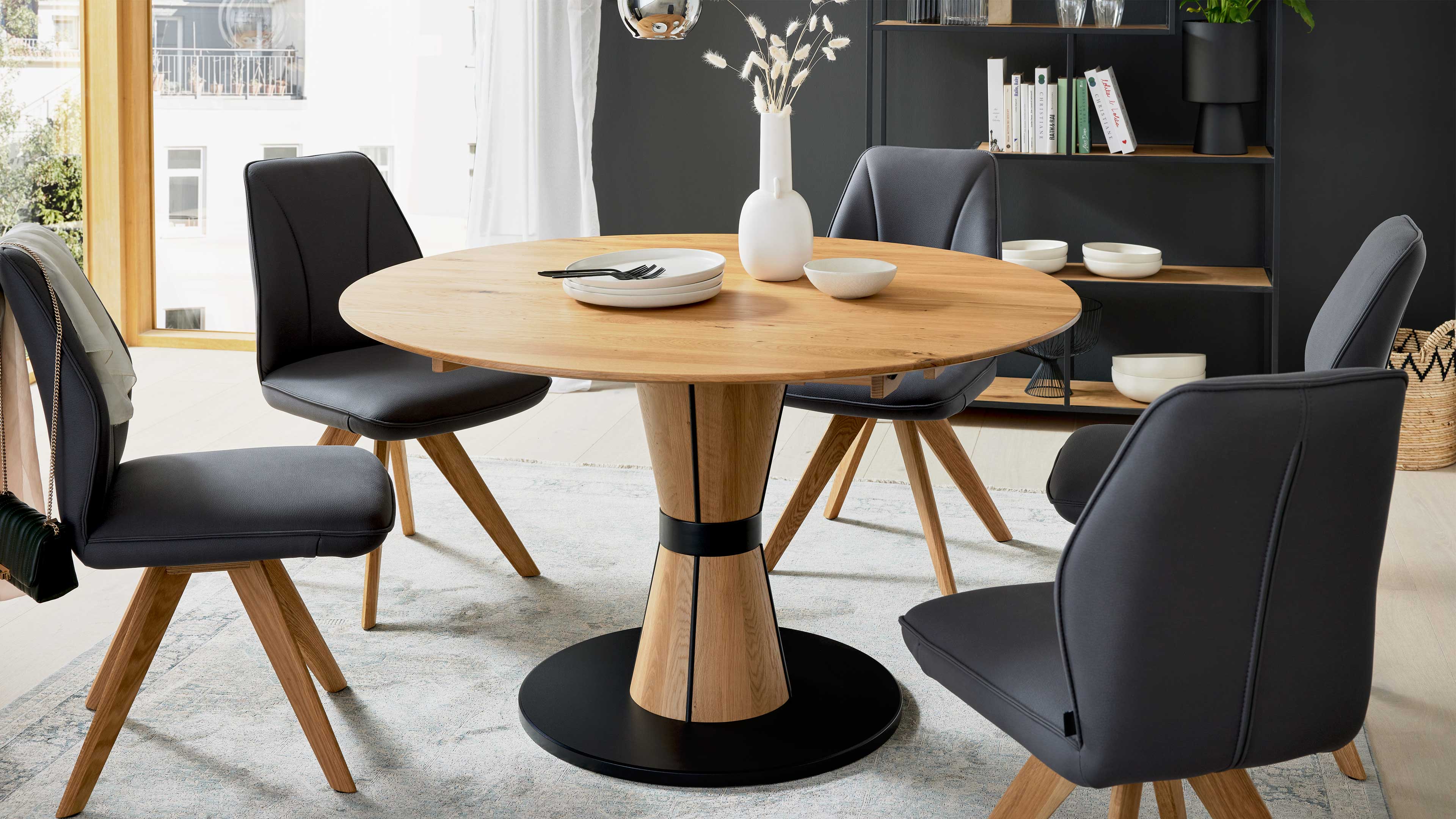 Runder Holztisch mit konischem Mittelfuß und schwarzer Basis, umgeben von vier grauen, gepolsterten Stühlen, mit Vase, Tellern und Besteck.