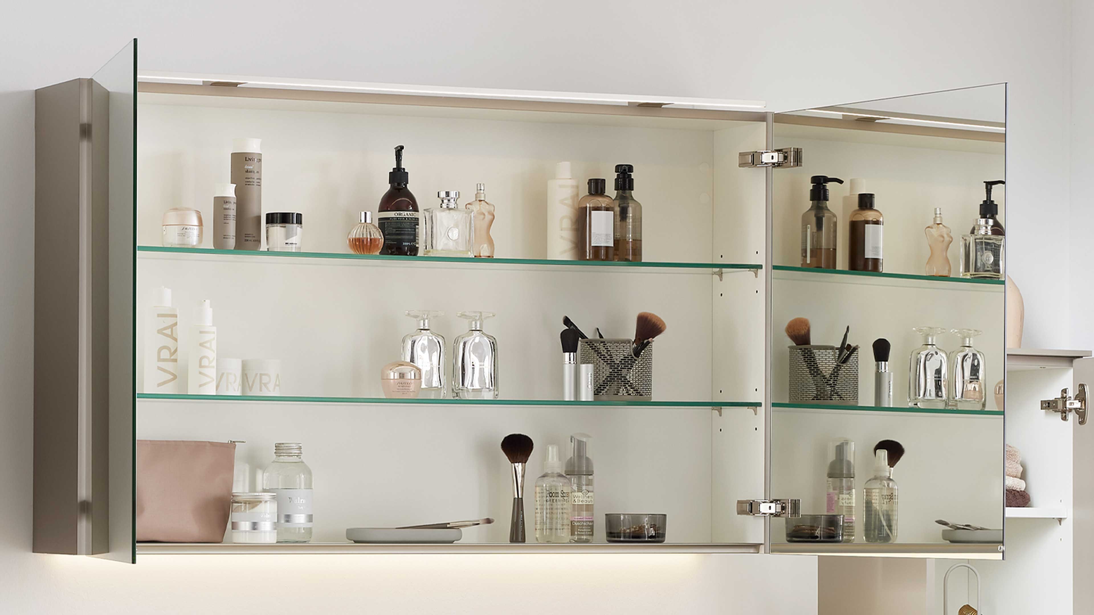 Wandhängender Badezimmerschrank mit offenen Spiegeltüren und drei Glasböden, bestückt mit Flaschen, Tiegeln und Make-up-Pinseln.