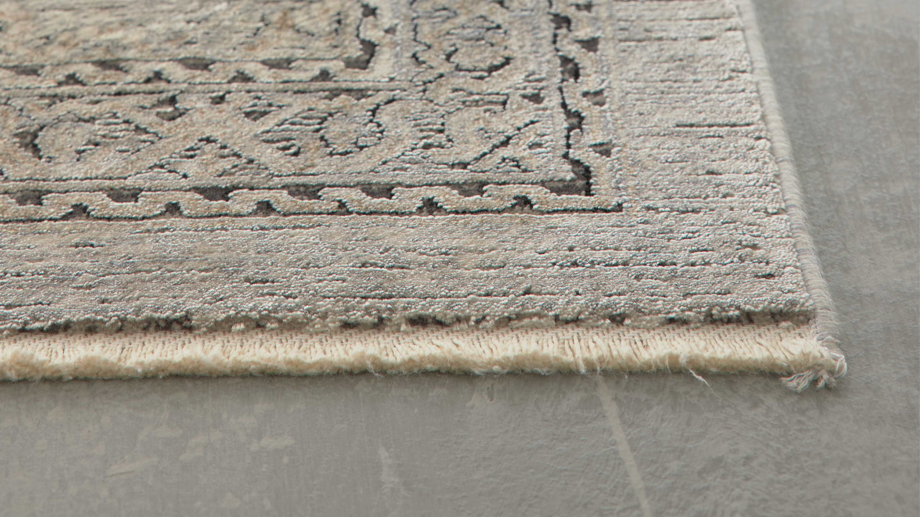 Rechte Ecke eines grau-beigen Teppichs mit ornamentaler Bordüre, sichtbarer Webkante und kurzem Fransenabschluss auf grauem Fliesenboden.