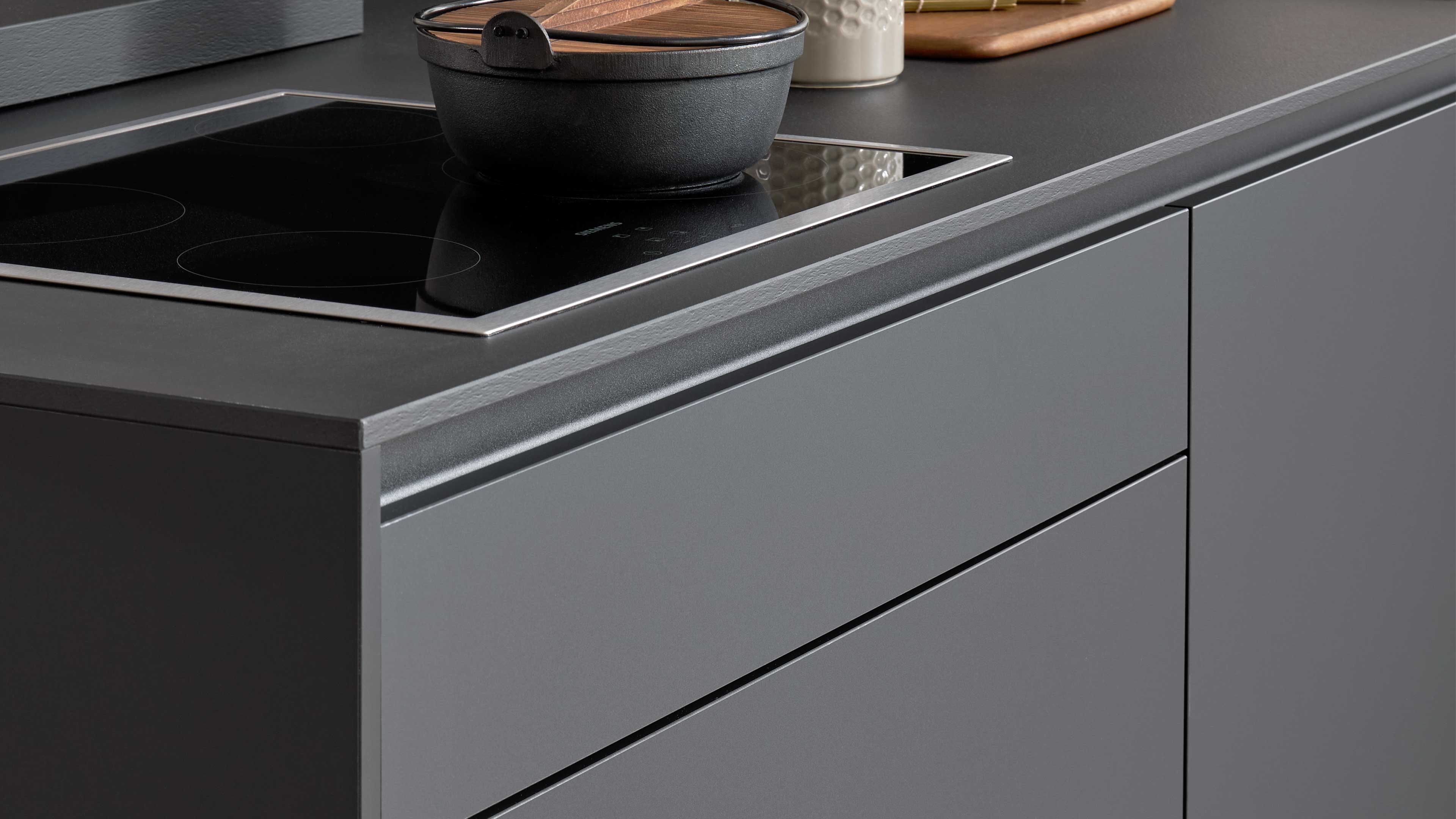 Graue Küchenfront mit integrierten Griffleisten, schwarzem Glaskeramik-Kochfeld und einem gusseisernen Topf auf der Arbeitsplatte.