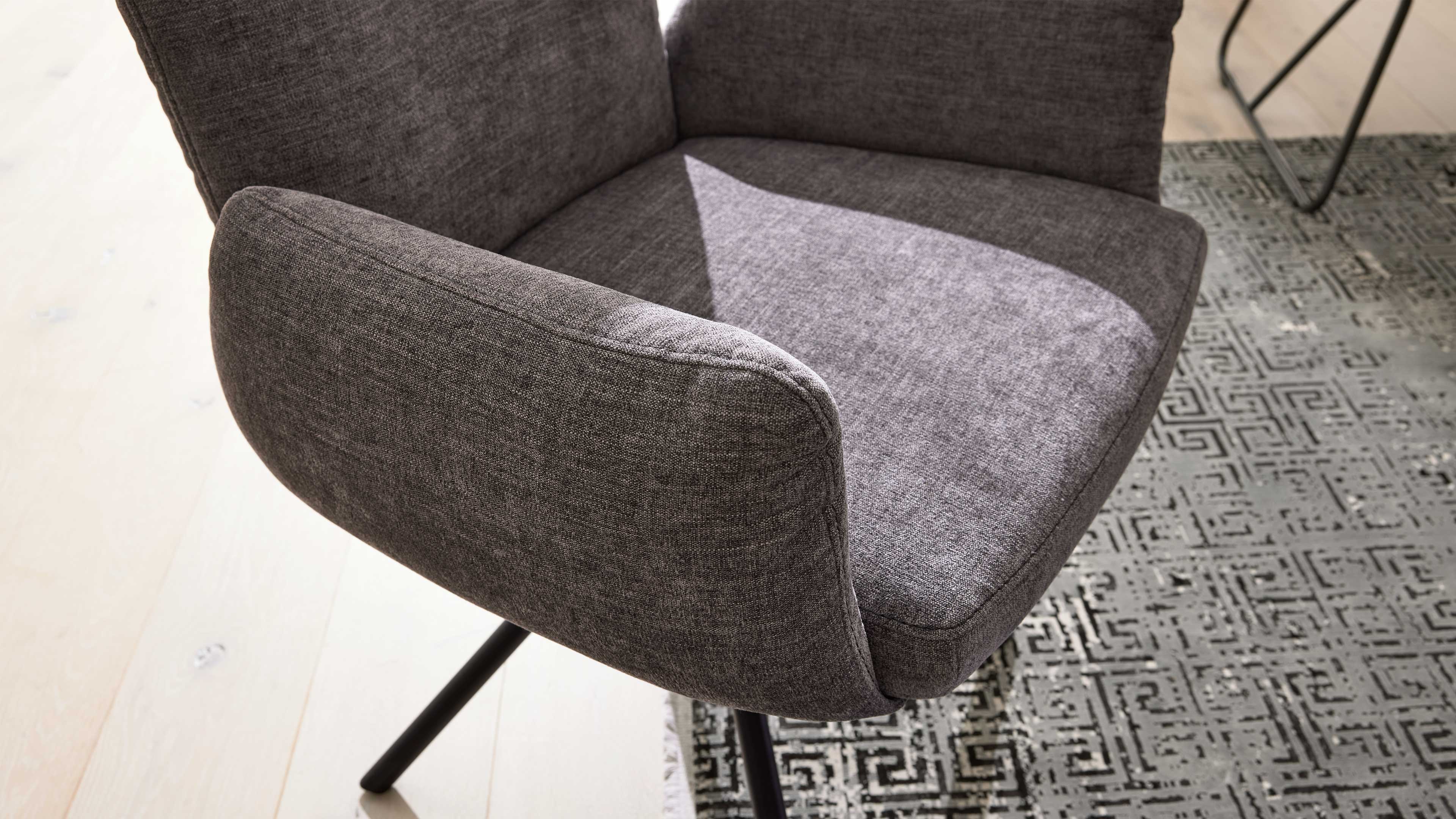 Grau bezogener Sessel mit gepolsterten Armlehnen und schwarzem Metallfuß, Sitz mit Sonnenfleck, auf hellem Boden und gemustertem Teppich.