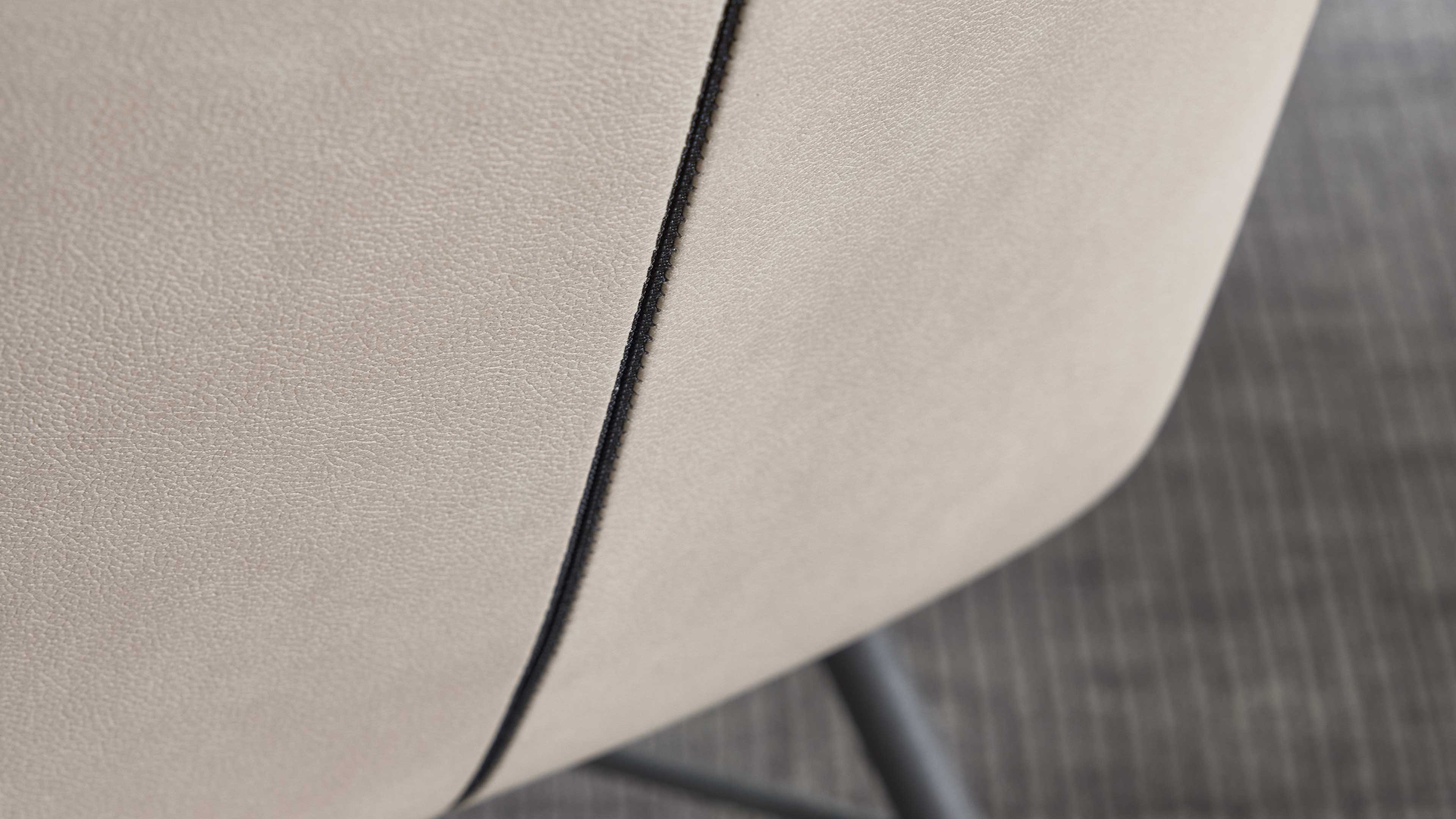 Beige Polsterfläche mit sichtbarer schwarzer Naht in der Mitte, rechts abgerundete Kante und schwarzes Metallbein.