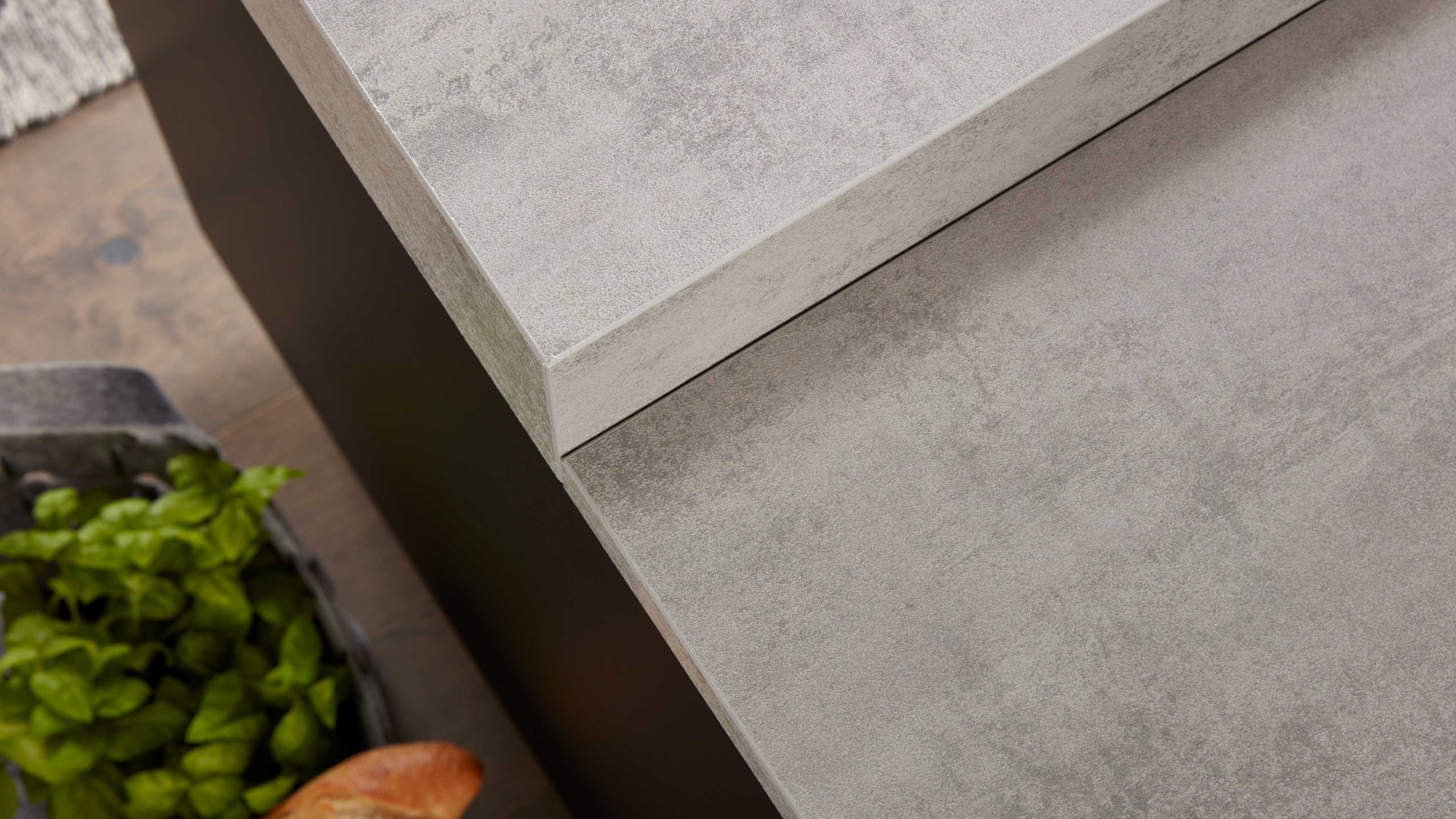 Eckige Arbeitsplatte mit grauer Betonoptik, sichtbare Kante und schmale Fuge, links Pflanzkübel mit grünen Kräutern und unten Brötchen.