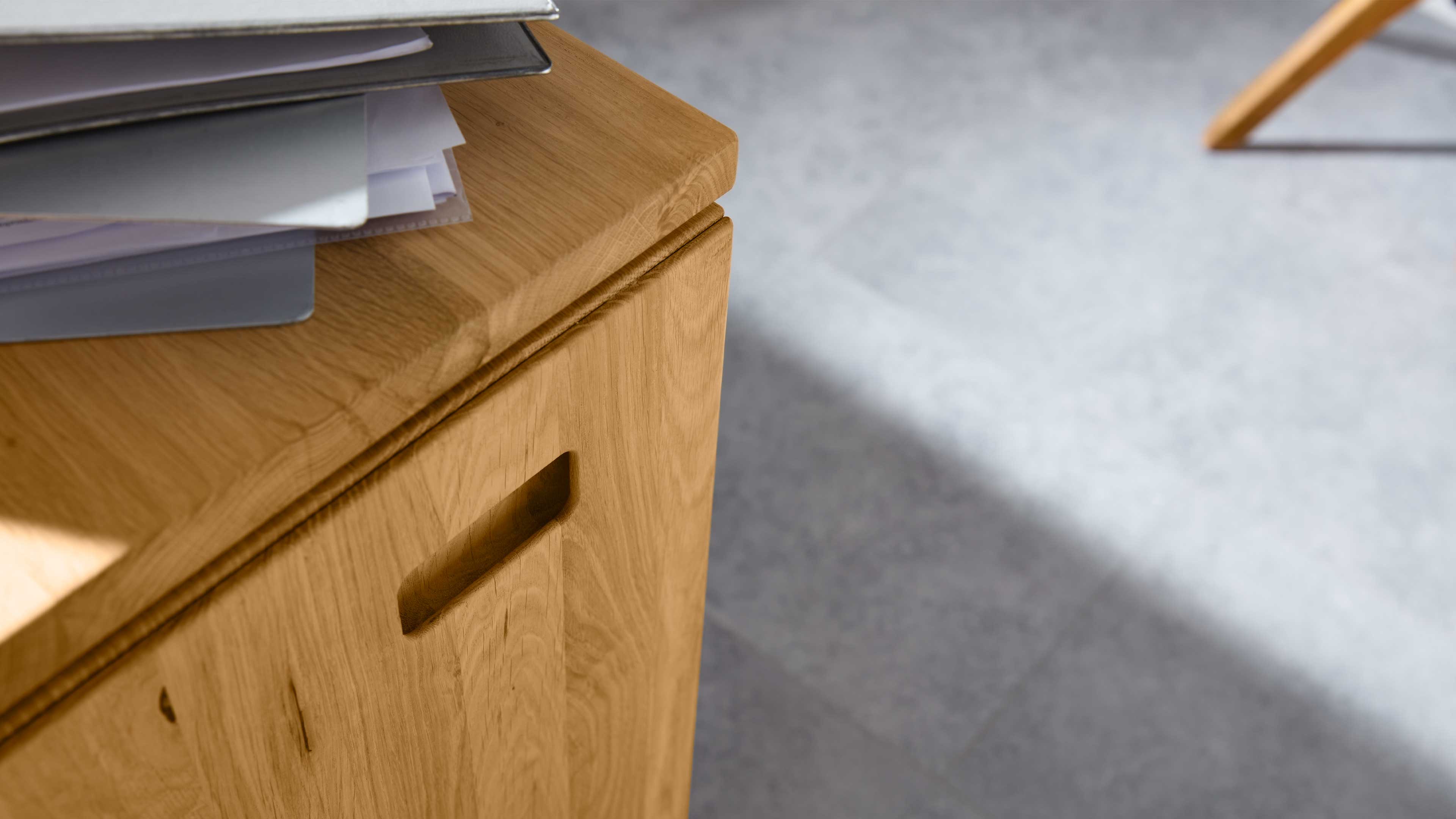 Eckansicht einer hellen Holzkommode mit eingearbeitetem Griff, lose Unterlagen auf der Oberseite und hellgrauem Boden.