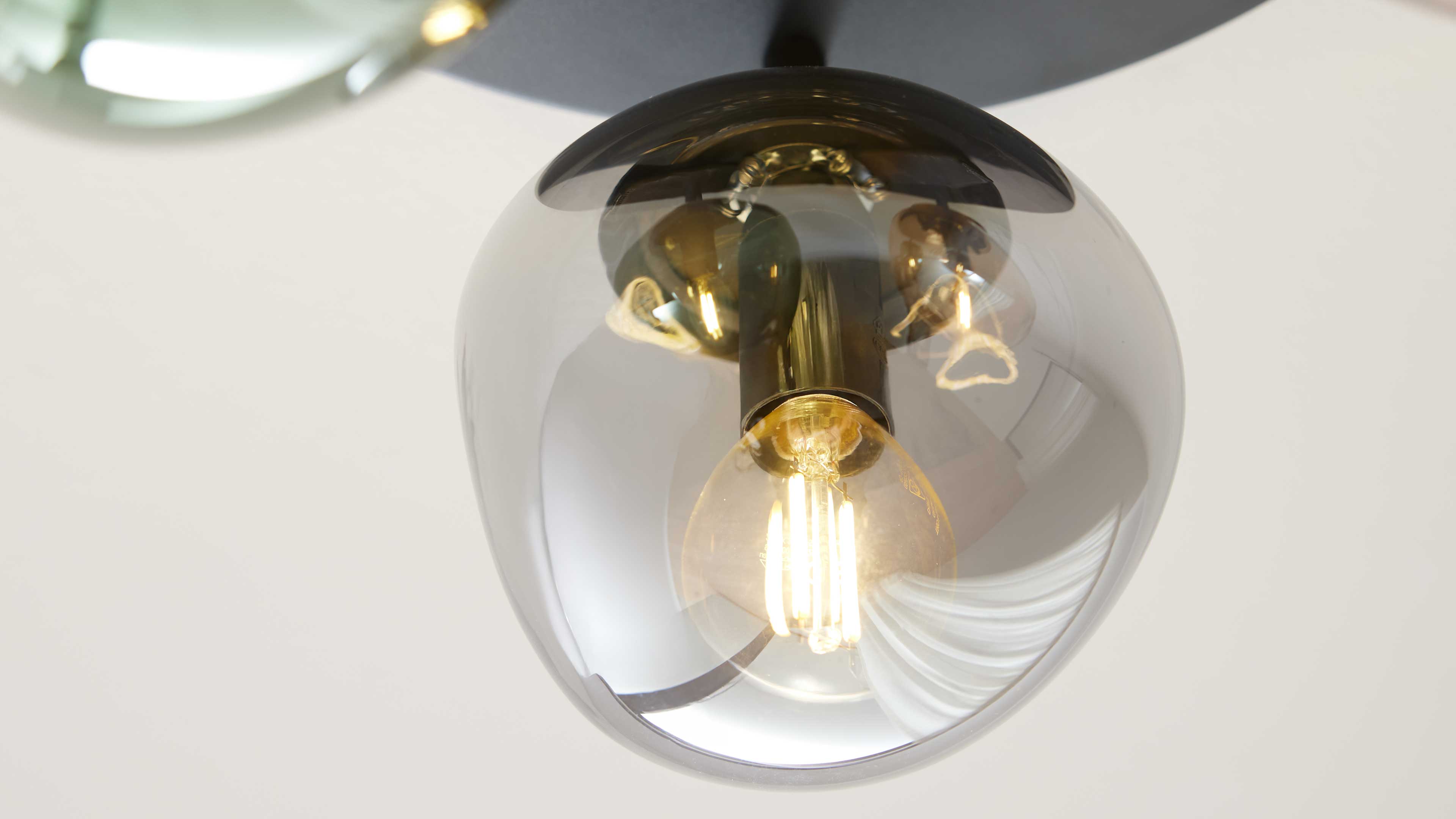 Deckenleuchte mit rauchig-transparentem Glasballon, sichtbarer warm leuchtender Filament-Glühbirne und goldfarbenem Fassungsdetail.