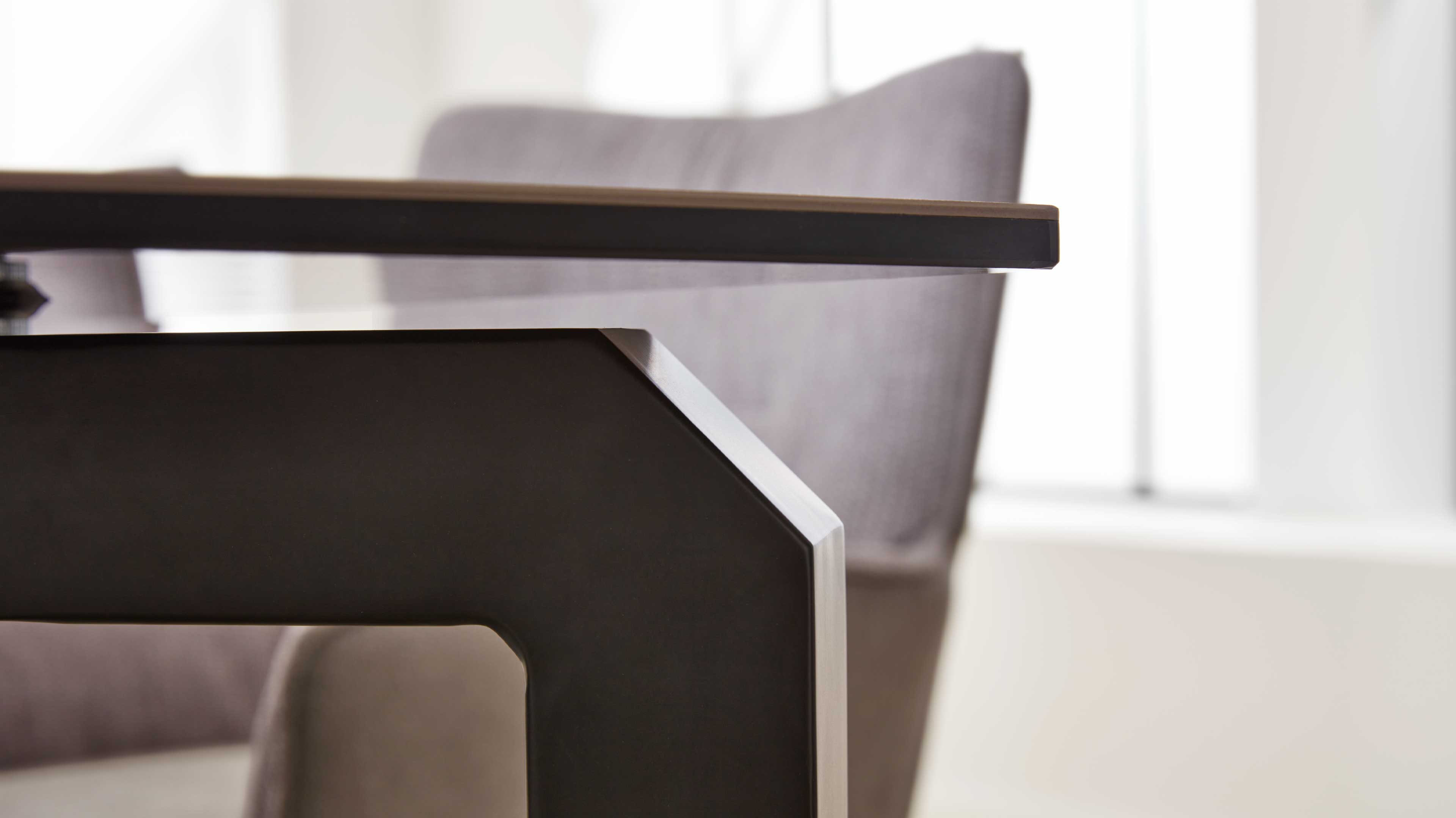 Nahaufnahme einer dunklen Metall-Tischbeinform mit abgeschrägter Kante und Holztischplatte, unscharfer Sessel und Fenster im Hintergrund.