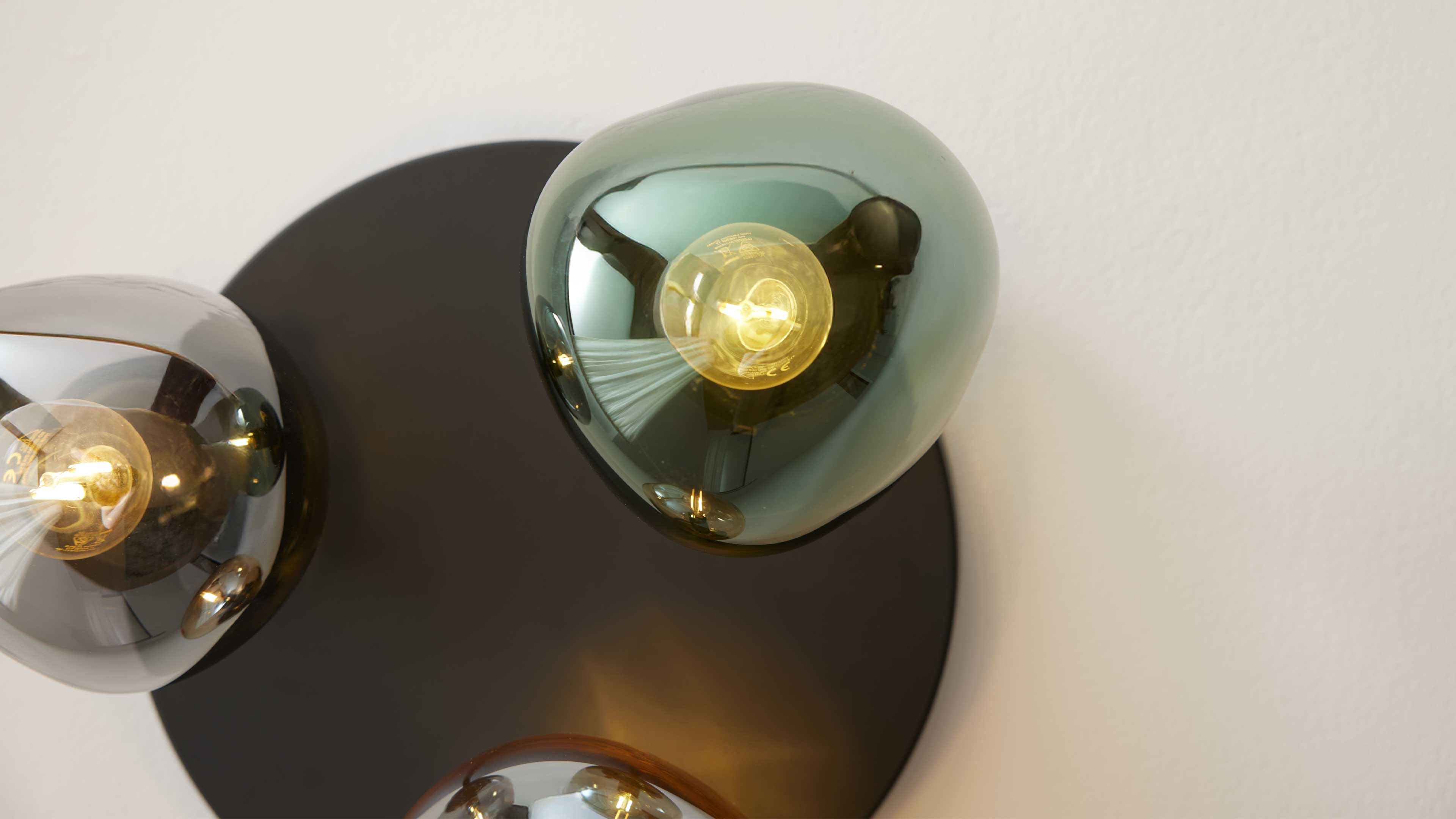 Wandlampe mit schwarzer runder Rückplatte und drei organischen, transparenten Glasschirmen in Grün/Grau mit eingeschalteten Birnen.