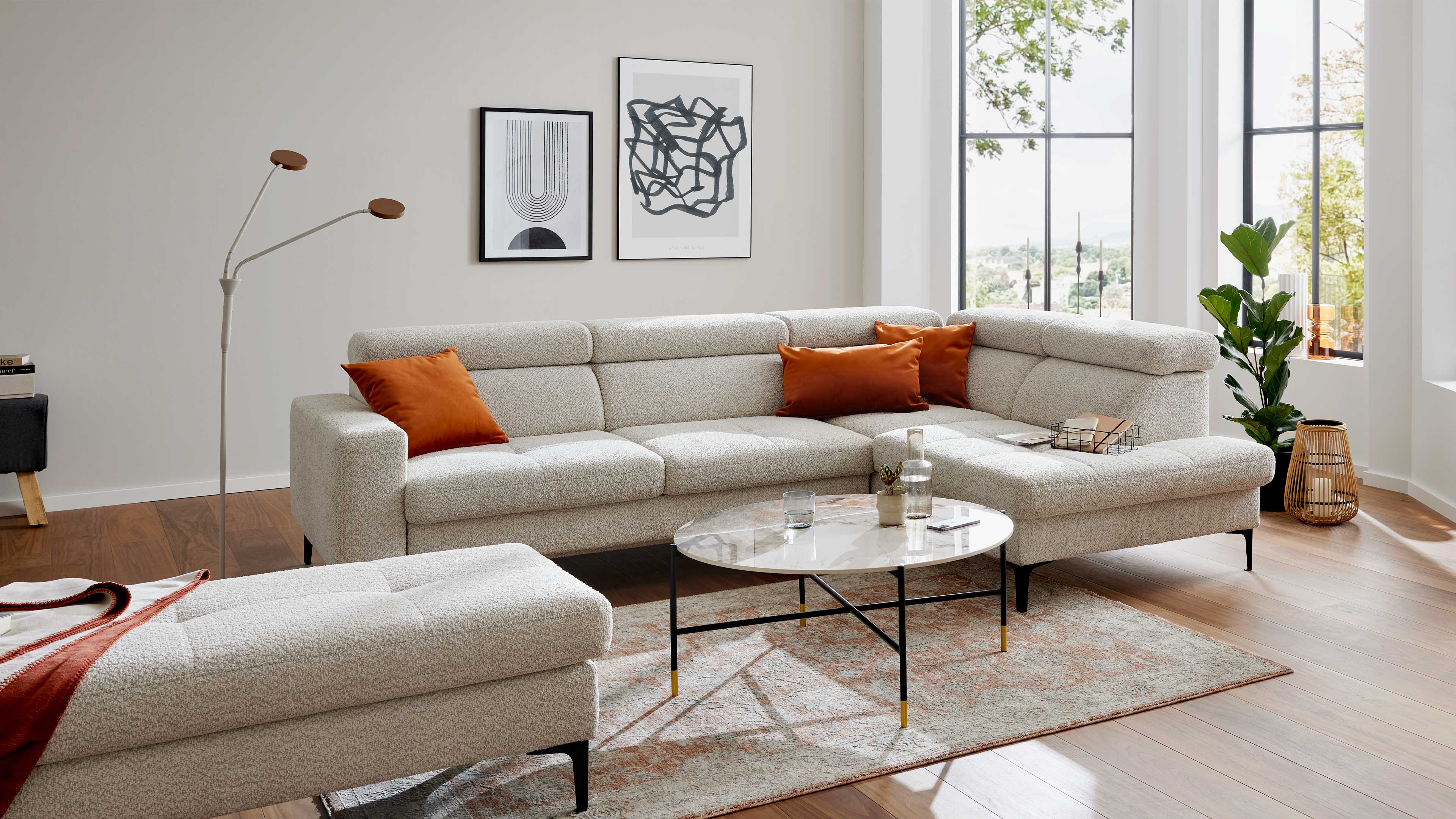 Helles L-förmiges Ecksofa mit drei orange Kissen, davor runder Couchtisch auf Teppich, links Stehleuchte, rechts Fenster und Pflanze.