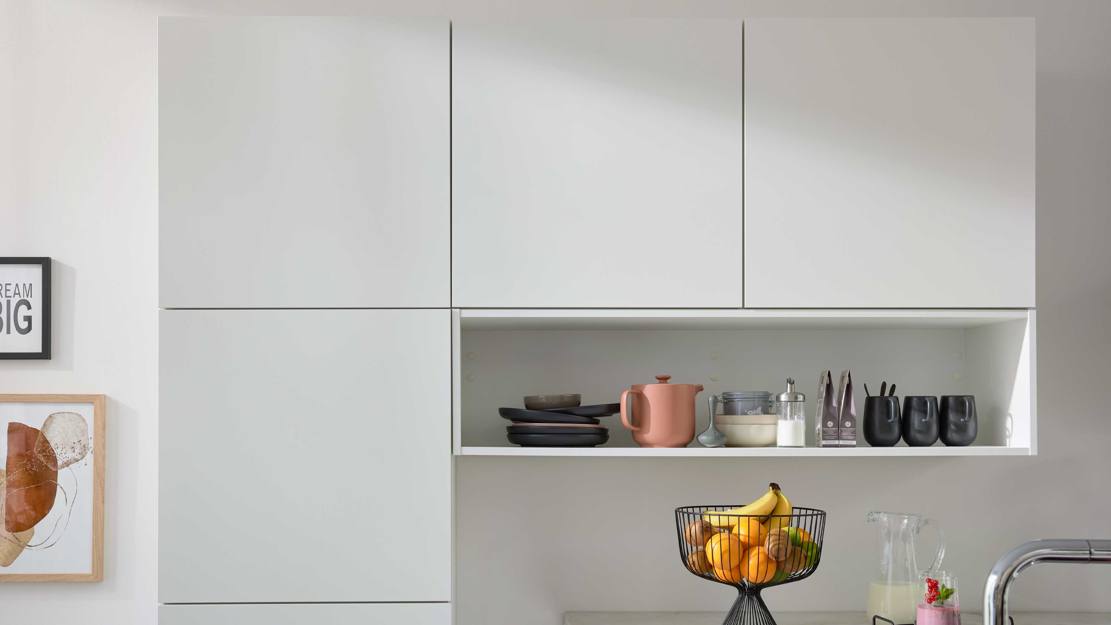 Weißer Wandhängeschrank mit offenem Fach, darin gestapelte Teller, rosafarbene Teekanne, Vorratsgläser und schwarze Tassen; darunter ein Obstkorb mit Bananen.