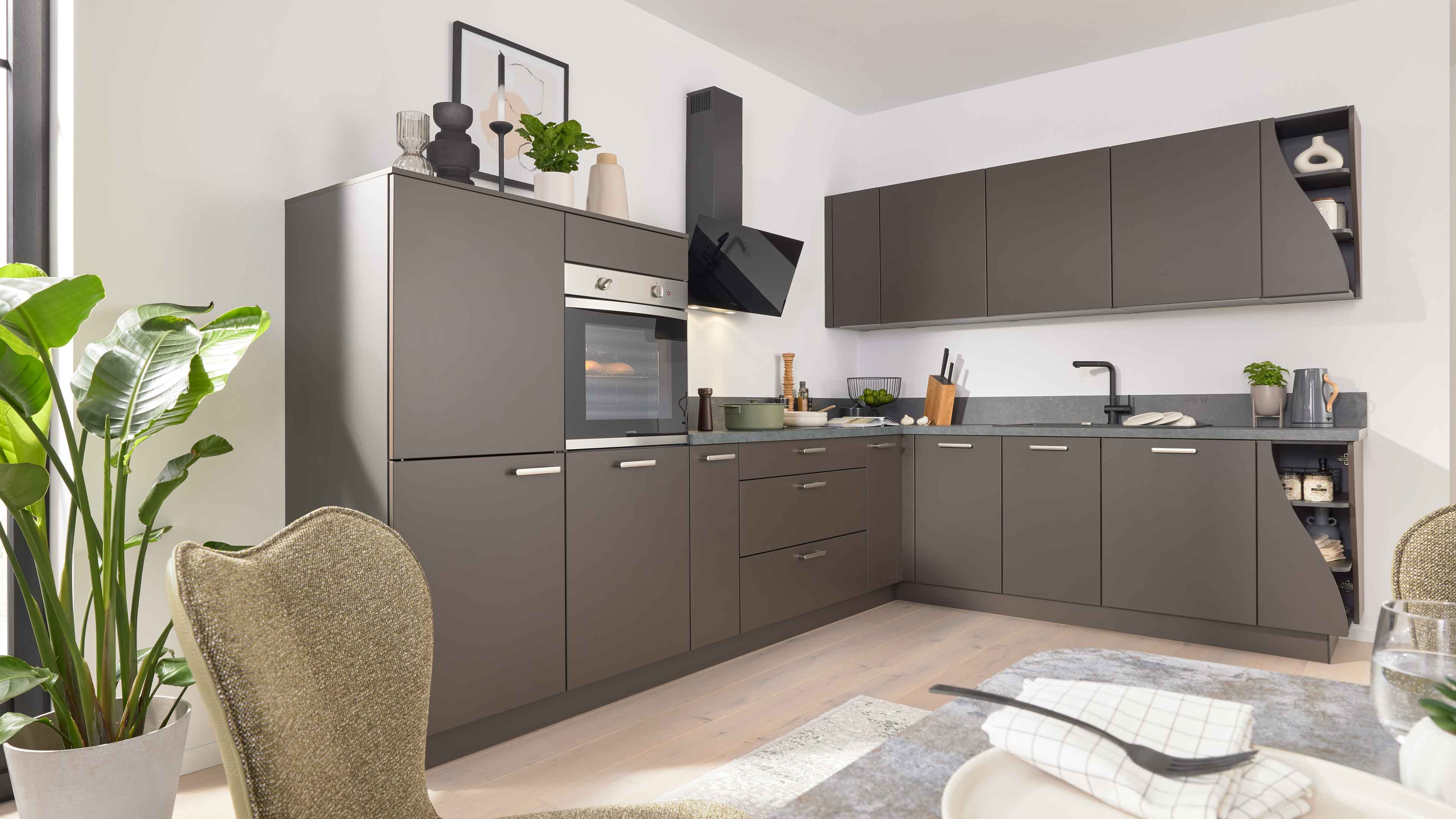 L-förmige dunkelgraue Küchenzeile mit eingebautem Ofen, schräger schwarzer Dunstabzugshaube, Spüle und Pflanzen.