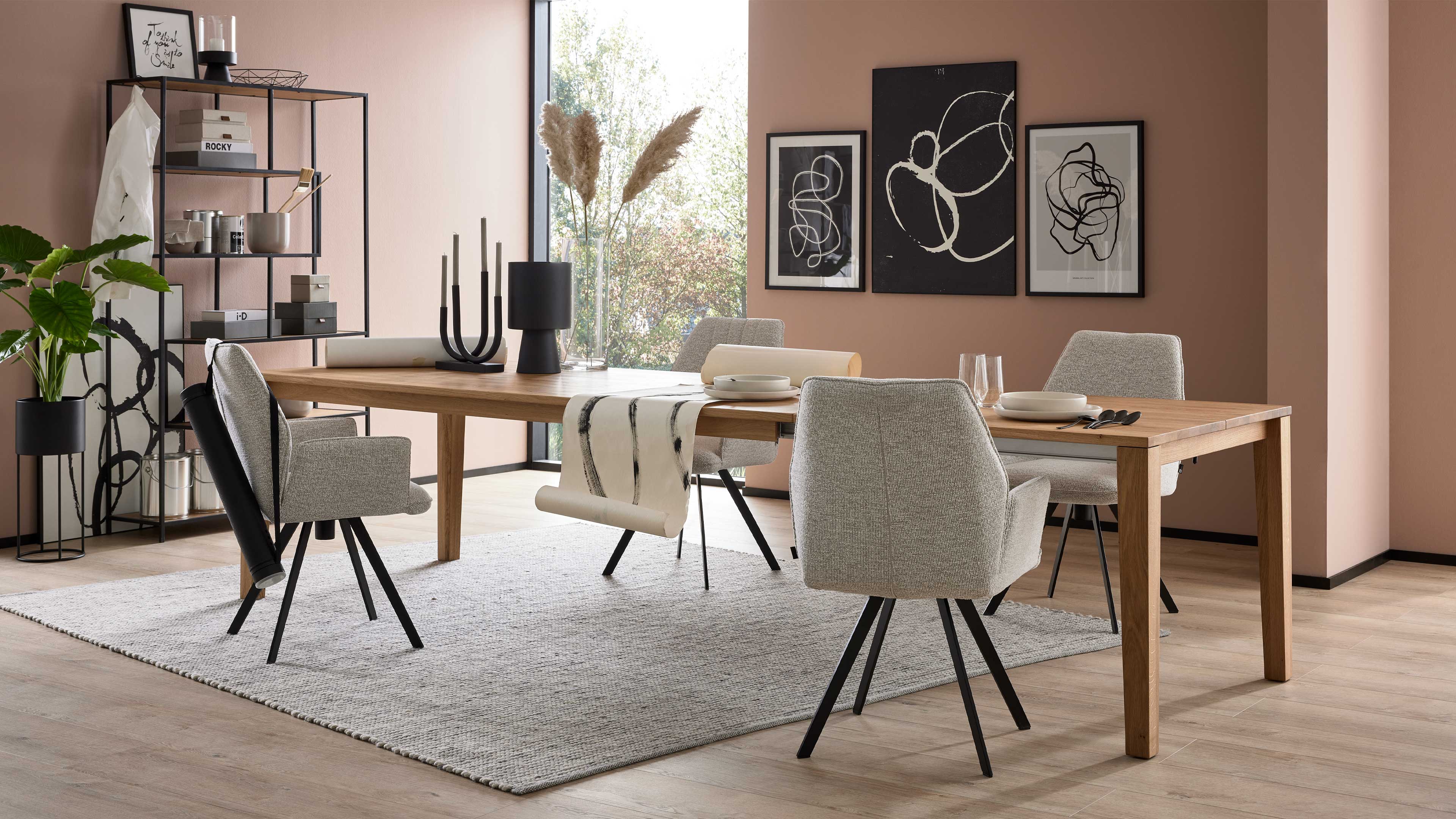 Rechteckiger Eichentisch mit vier grauen Stühlen auf hellem Teppich vor rosafarbenen Wänden, links Regal und Pflanzen.