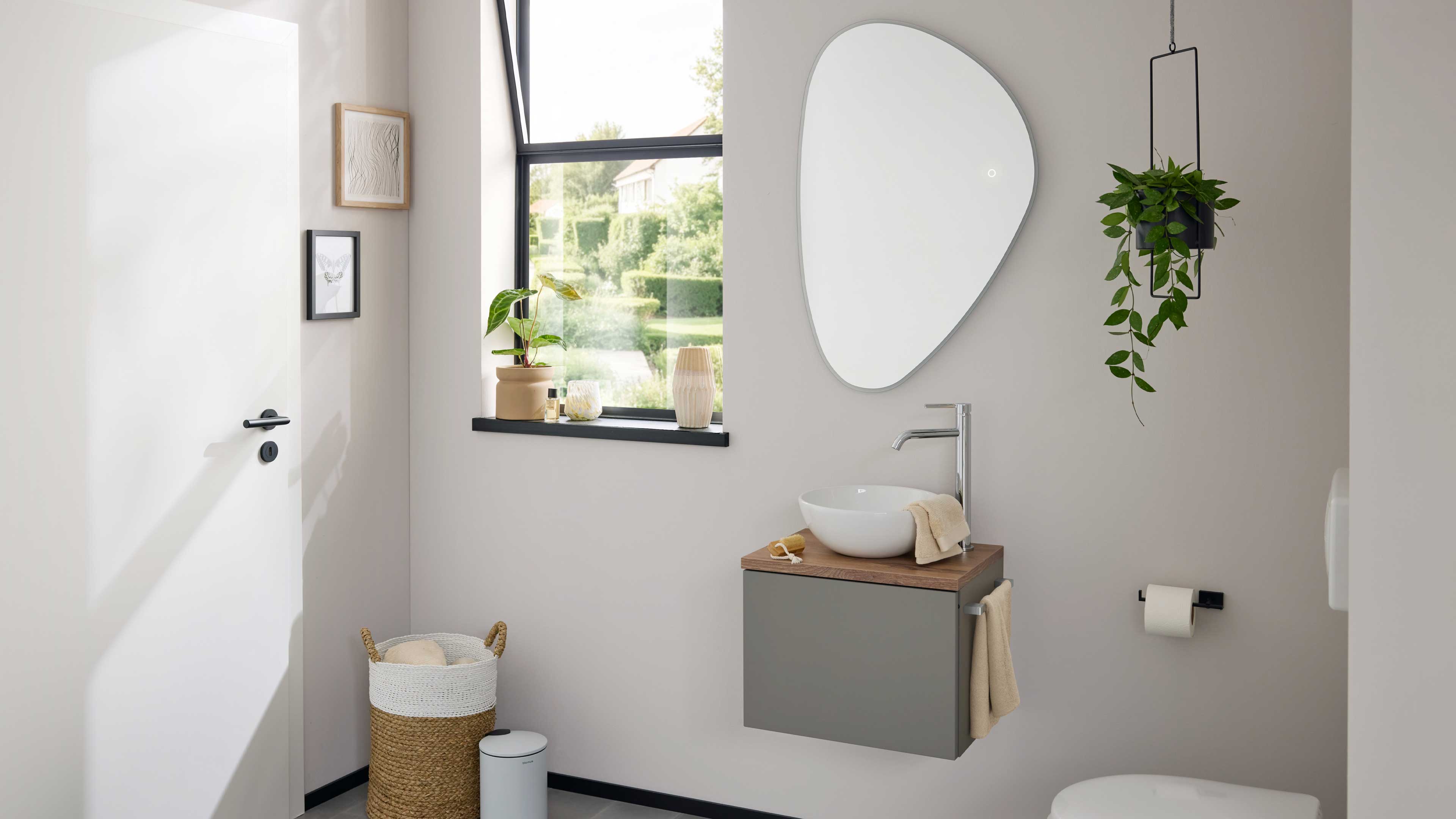Wandhängender grauer Waschplatz mit Holzplatte und rundem Aufsatzbecken unter asymmetrischem Spiegel, neben Fenster, Hängetopf und Korb.