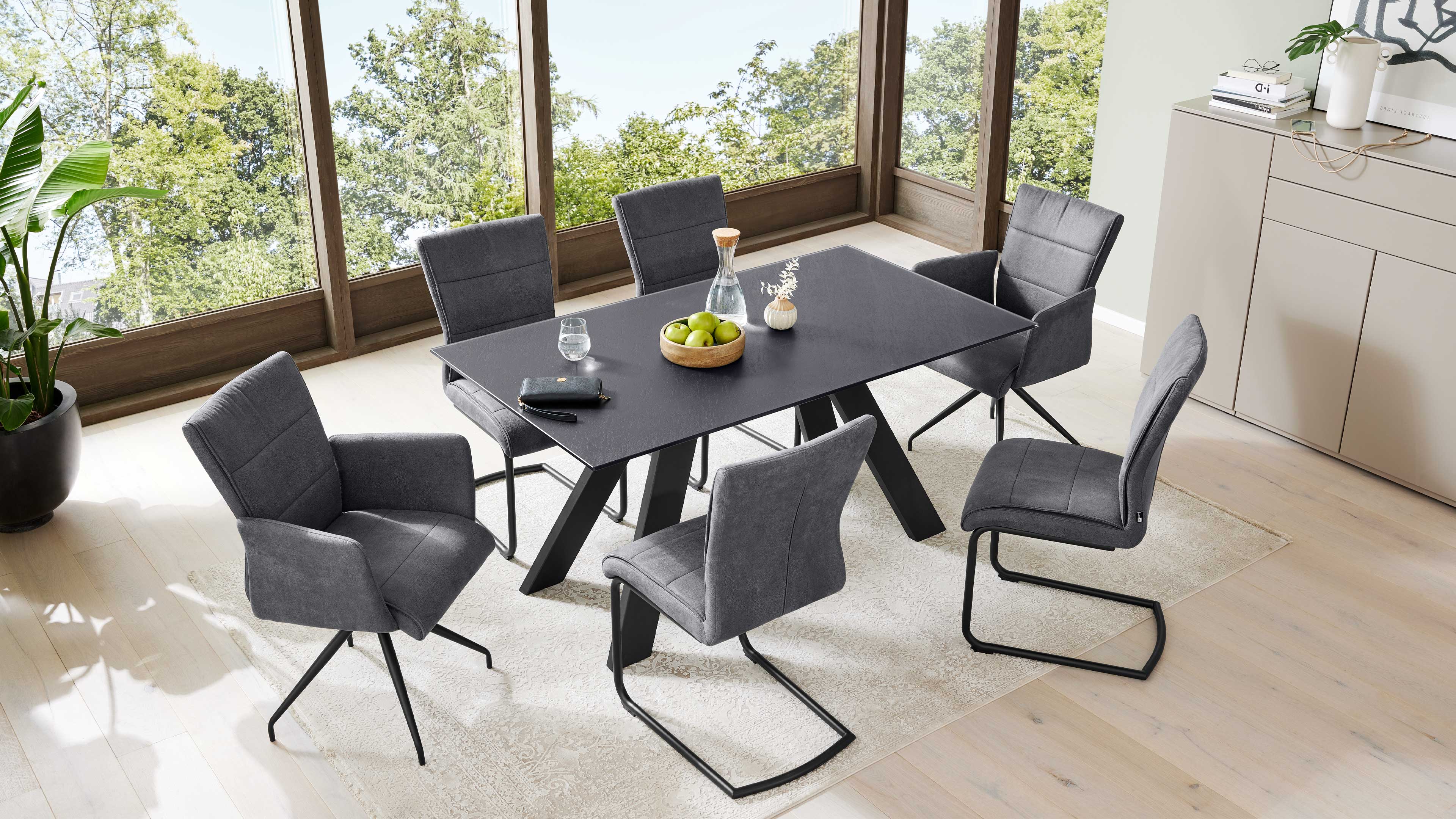 Schwarzer rechteckiger Esstisch mit sechs grauen, gepolsterten Stühlen, Holzschale mit grünen Äpfeln vor großen Fenstern.