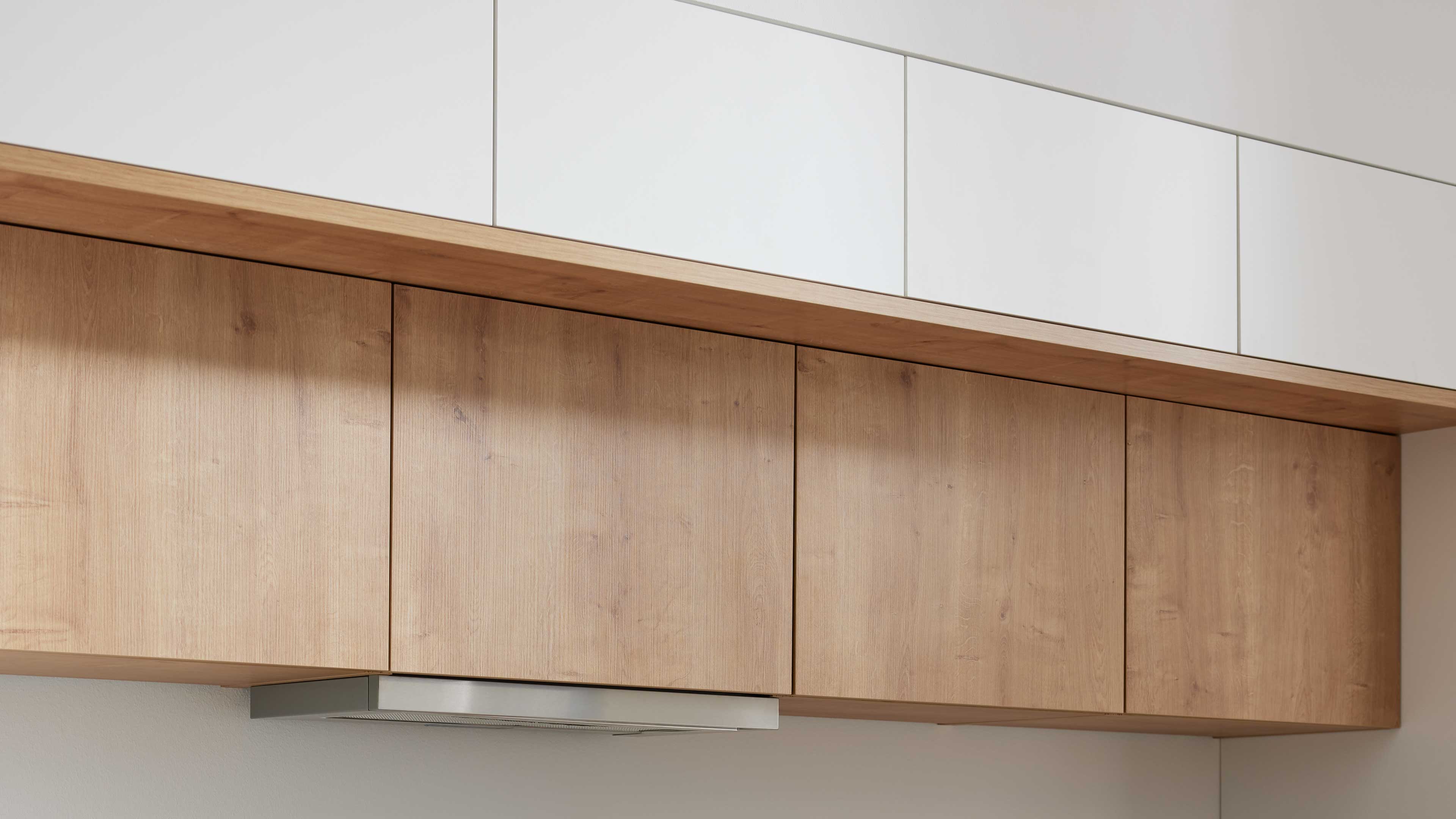 Untere Hängeschränke mit vier Holzfronten, darunter eine flache Edelstahlhaube, darüber eine Holzablage und weiße Oberschränke.