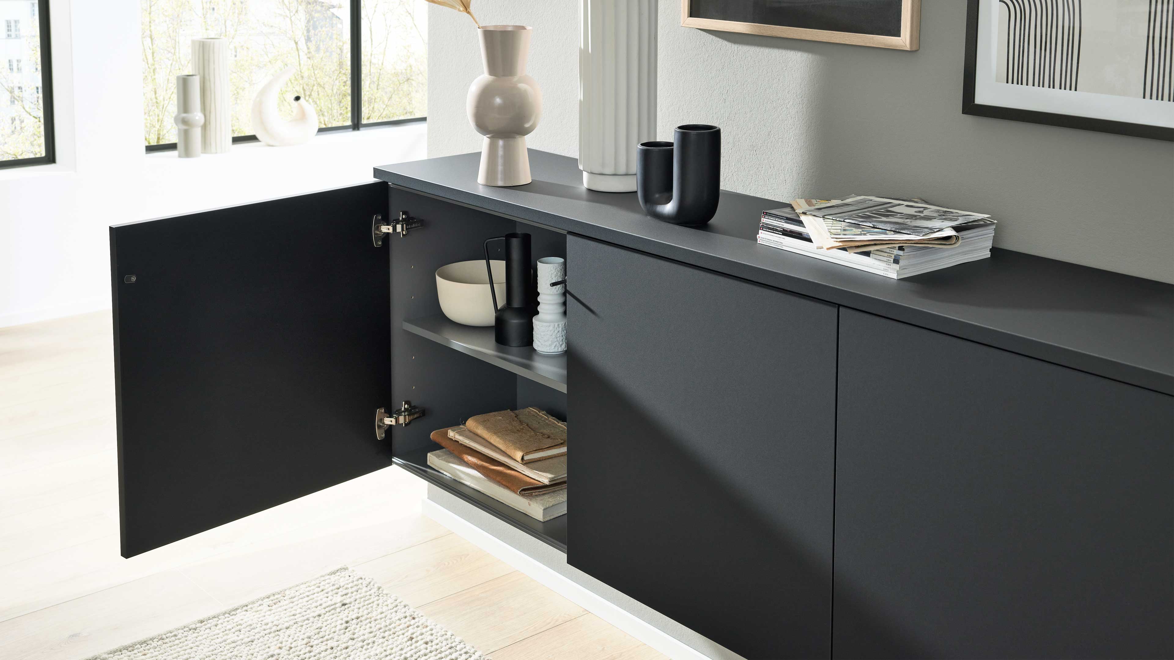 Schwarzes wandhängendes Sideboard mit offener linker Tür und zwei Regalfächern mit Schalen, Vasen und Büchern.