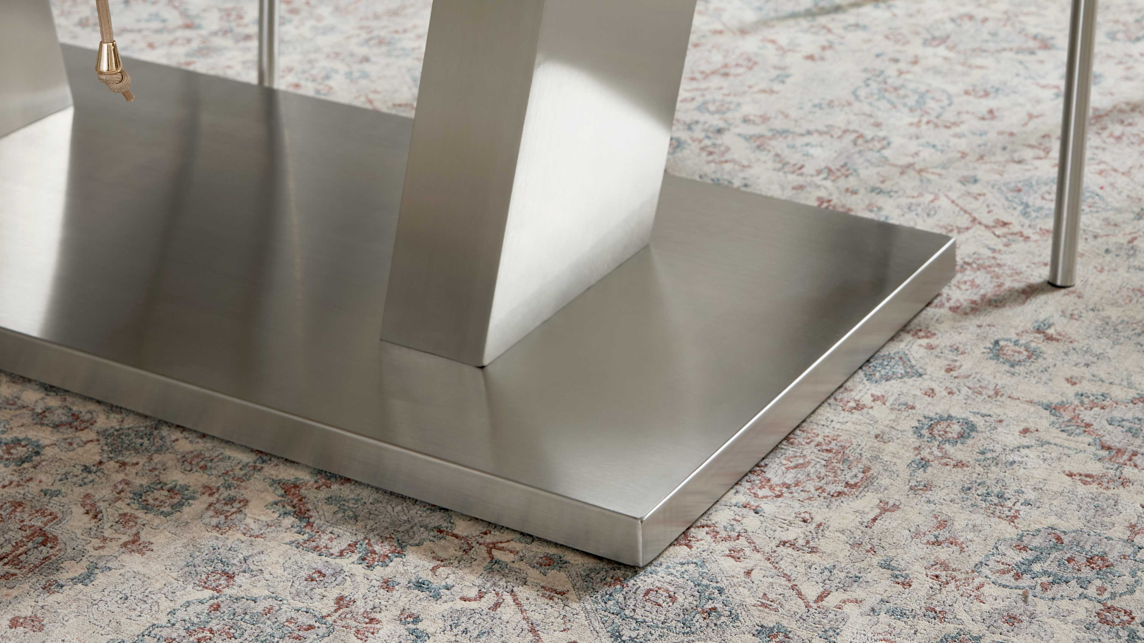 Rechteckige metallische Tischplatte mit zentralem abgeschrägtem Stützpfeiler und dünnem Rundfuß rechts auf gemustertem Teppich.