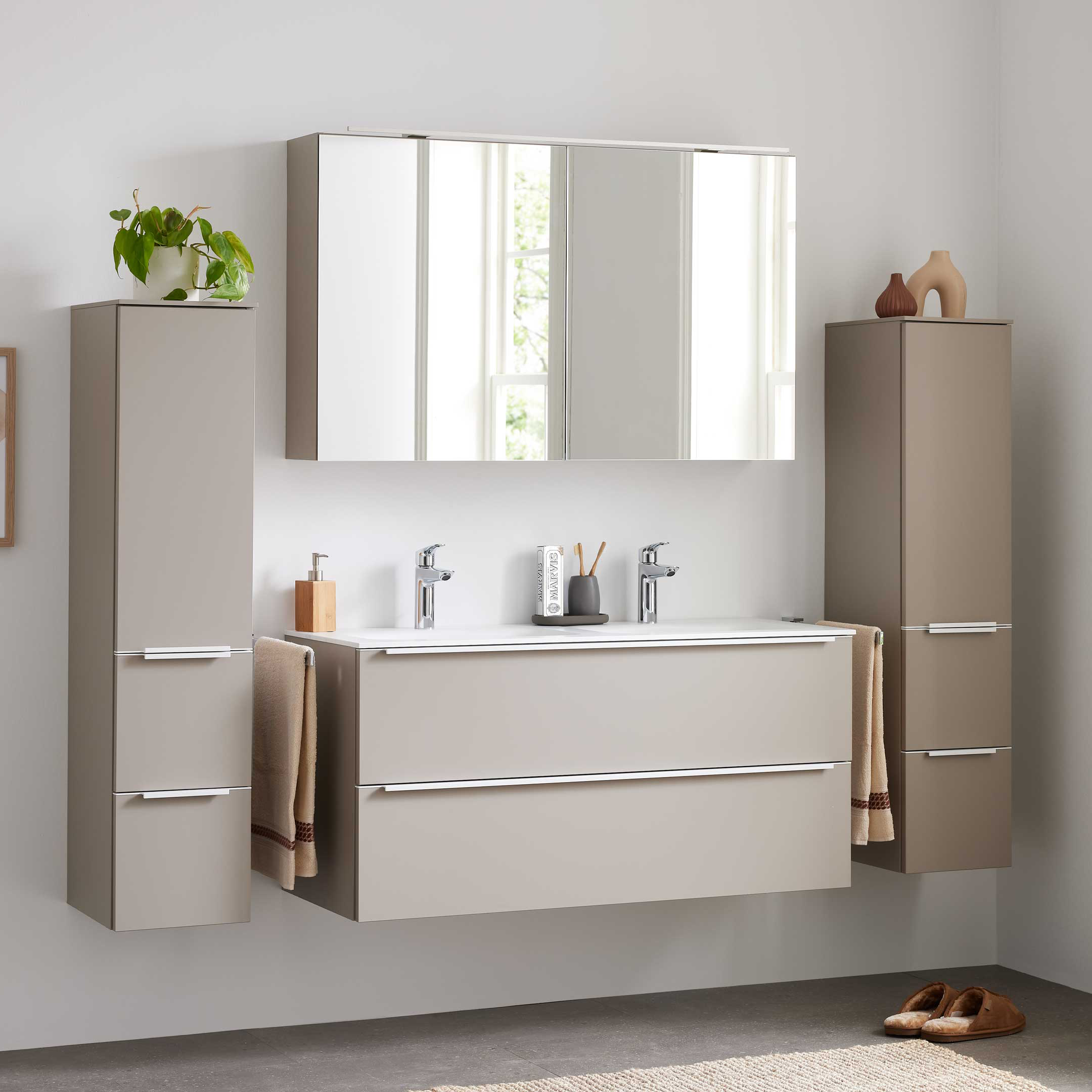 Wandhängende beige Badezimmerschränke mit Doppelwaschtisch, Spiegelschrank, zwei hohen Seitenschränken und Handtüchern.