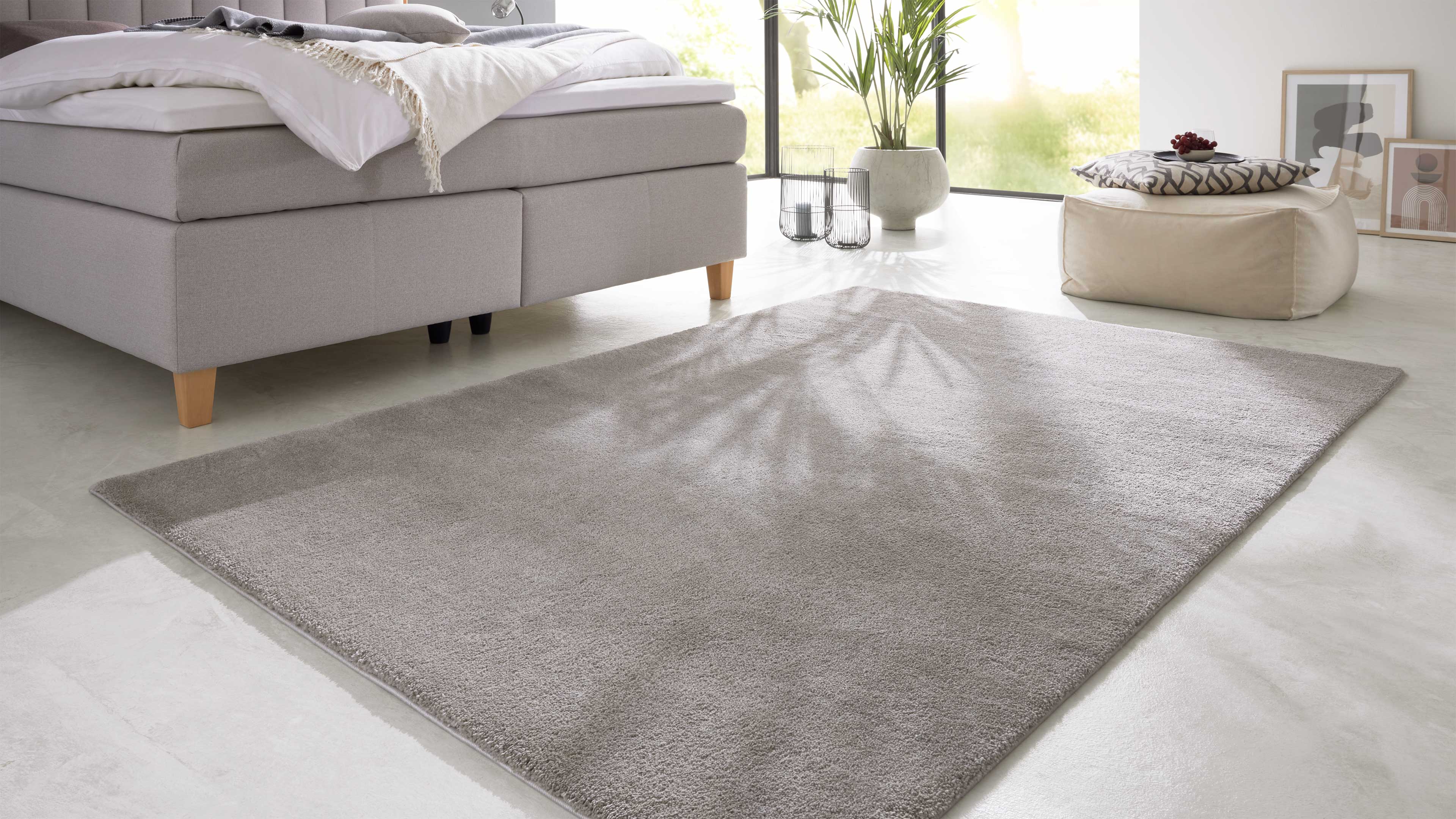 Großes hellgraues Rechteck-Teppich liegt vor einem hellgrauen Polsterbett, rechts Pouf, Pflanze und Bilder.