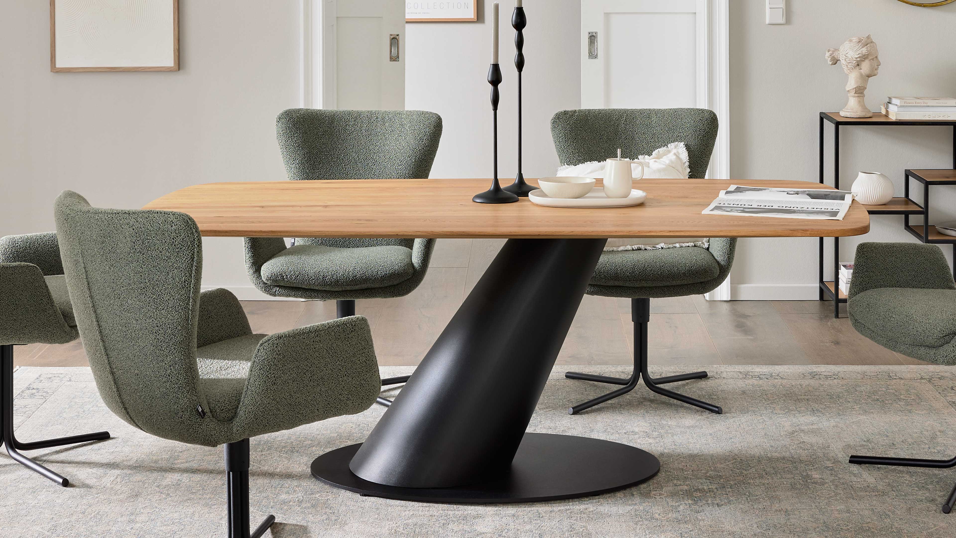 Tisch mit hellbrauner Holzplatte auf schrägem schwarzem Fuß, umgeben von vier grünen Drehstühlen, auf der Platte Kerzen und Zeitung.