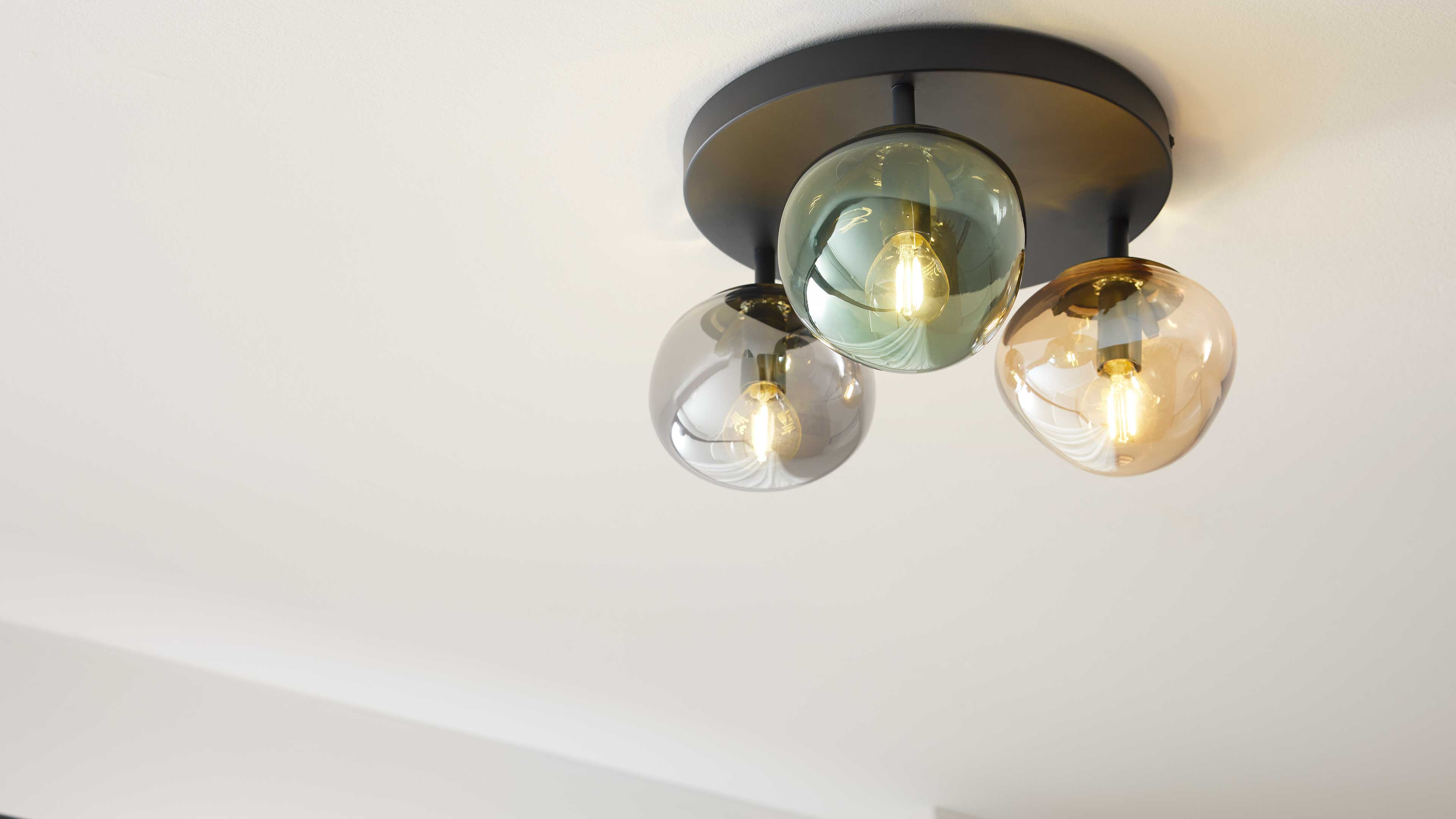Deckeuchte mit runder schwarzer Montageplatte und drei unterschiedlich getönten Glasglocken mit sichtbaren Leuchtmitteln.