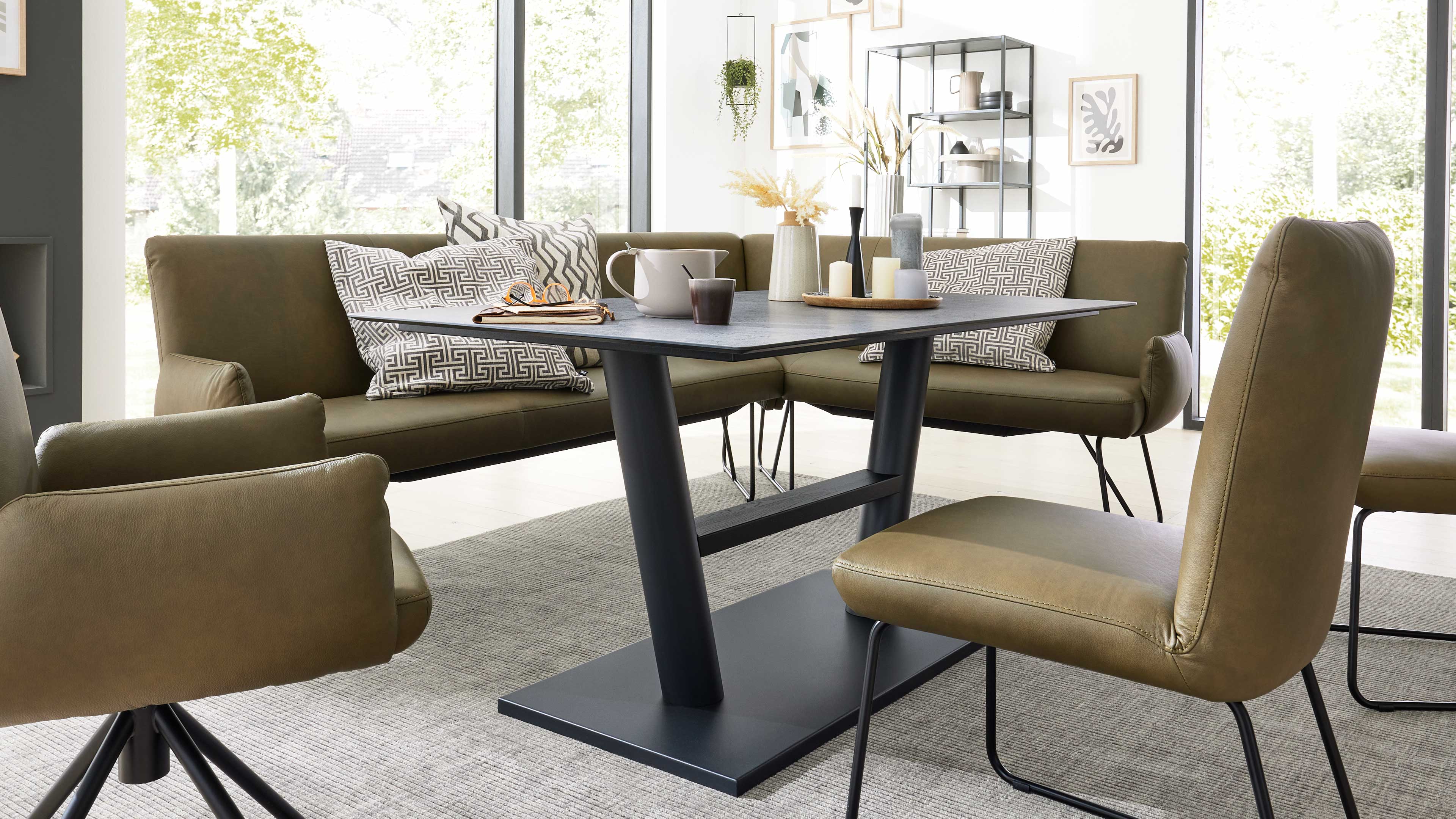 Rechteckiger schwarzer Esstisch mit zentraler Säulenbasis, umgeben von olivgrüner Eckbank und Stühlen, mit Kerzen, Vasen und Tassen.