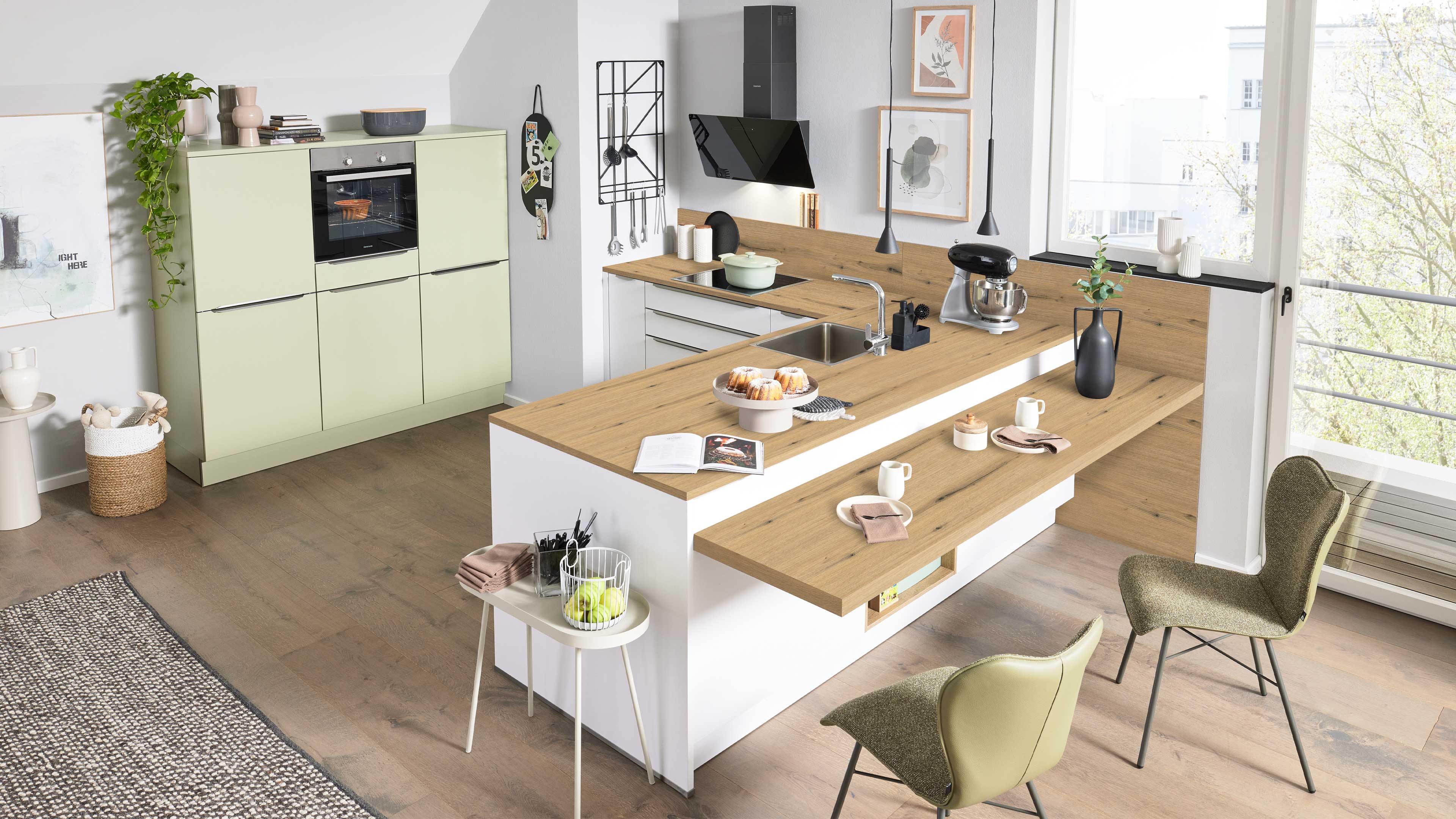Kücheninsel mit Holzarbeitsplatte, Spülbecken, Kuchen und Küchenmaschine, davor zwei grüne Stühle; links grüne Hochschränke mit Backofen.
