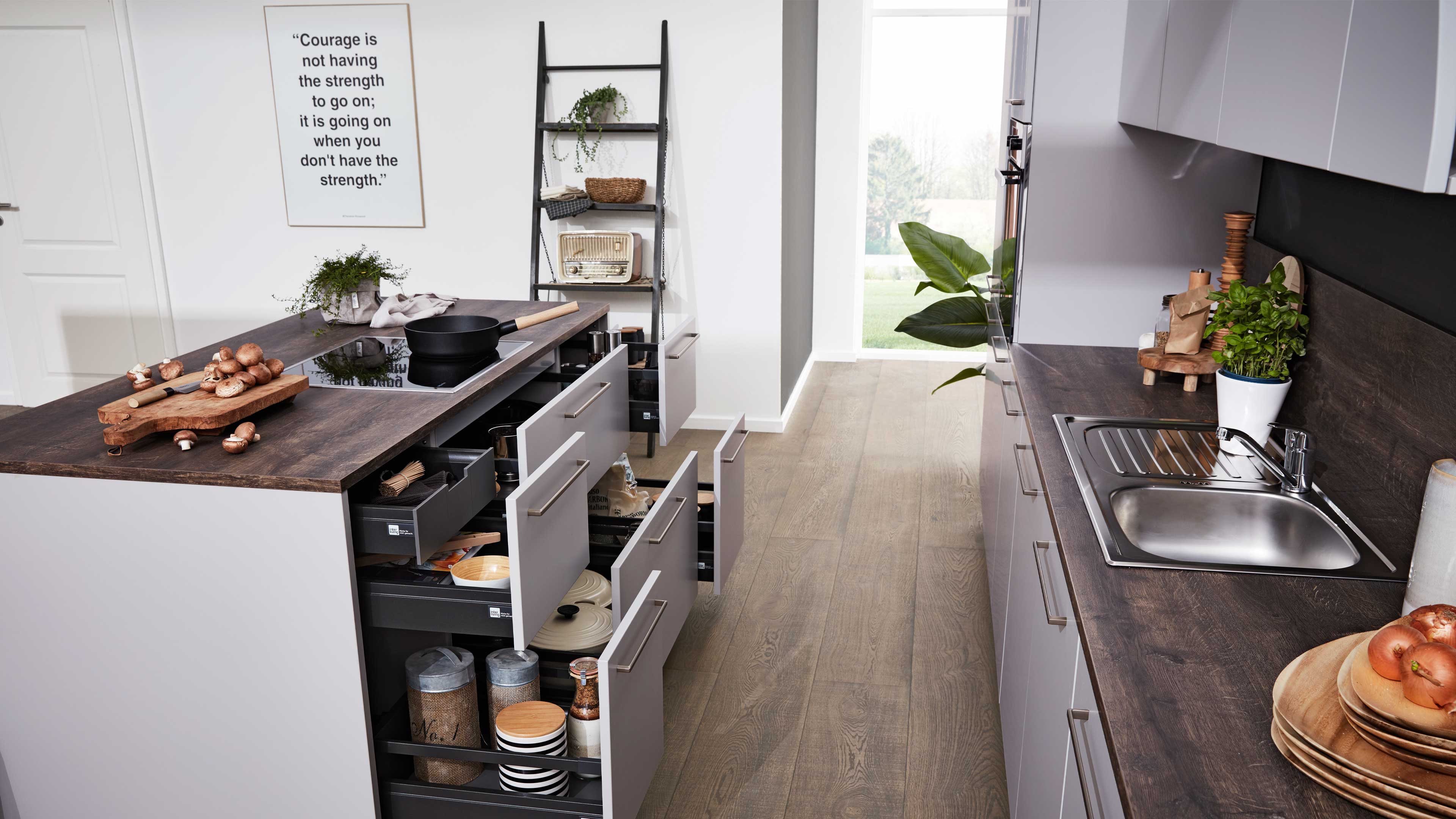 Kücheninsel mit dunkler Holzarbeitsplatte, geöffneten grauen Schubladen, Kochfeld und Schneidebrett mit Pilzen, rechts Spüle.