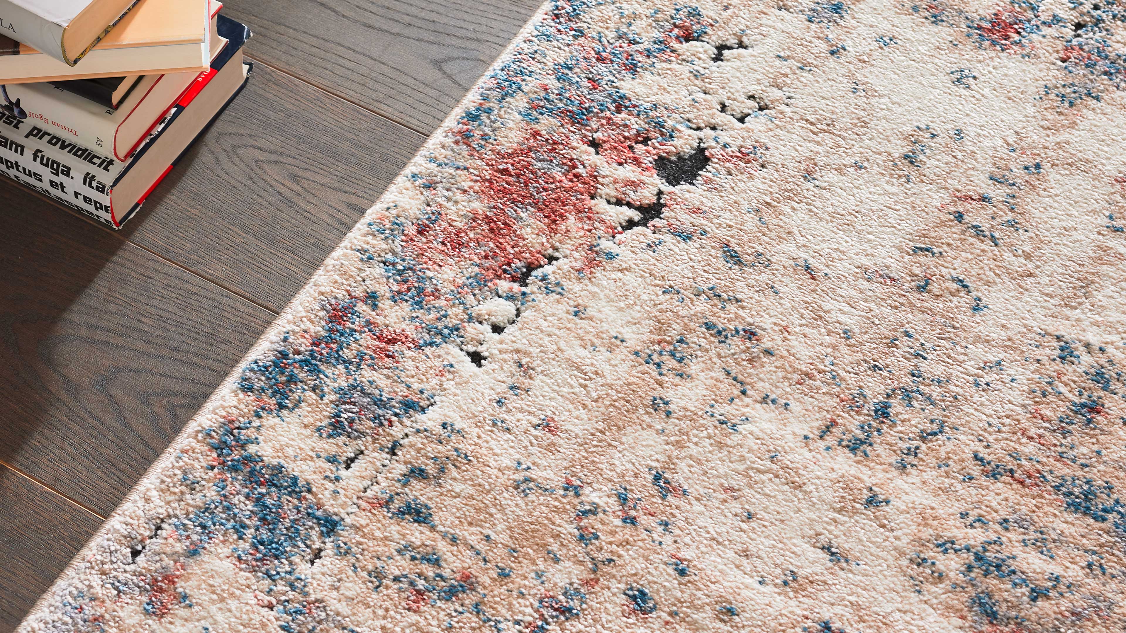 Beiger Teppich mit rot-blau-schwarzen Sprenkeln und unregelmäßigem Muster liegt auf dunklem Holzboden, links ein Bücherstapel.