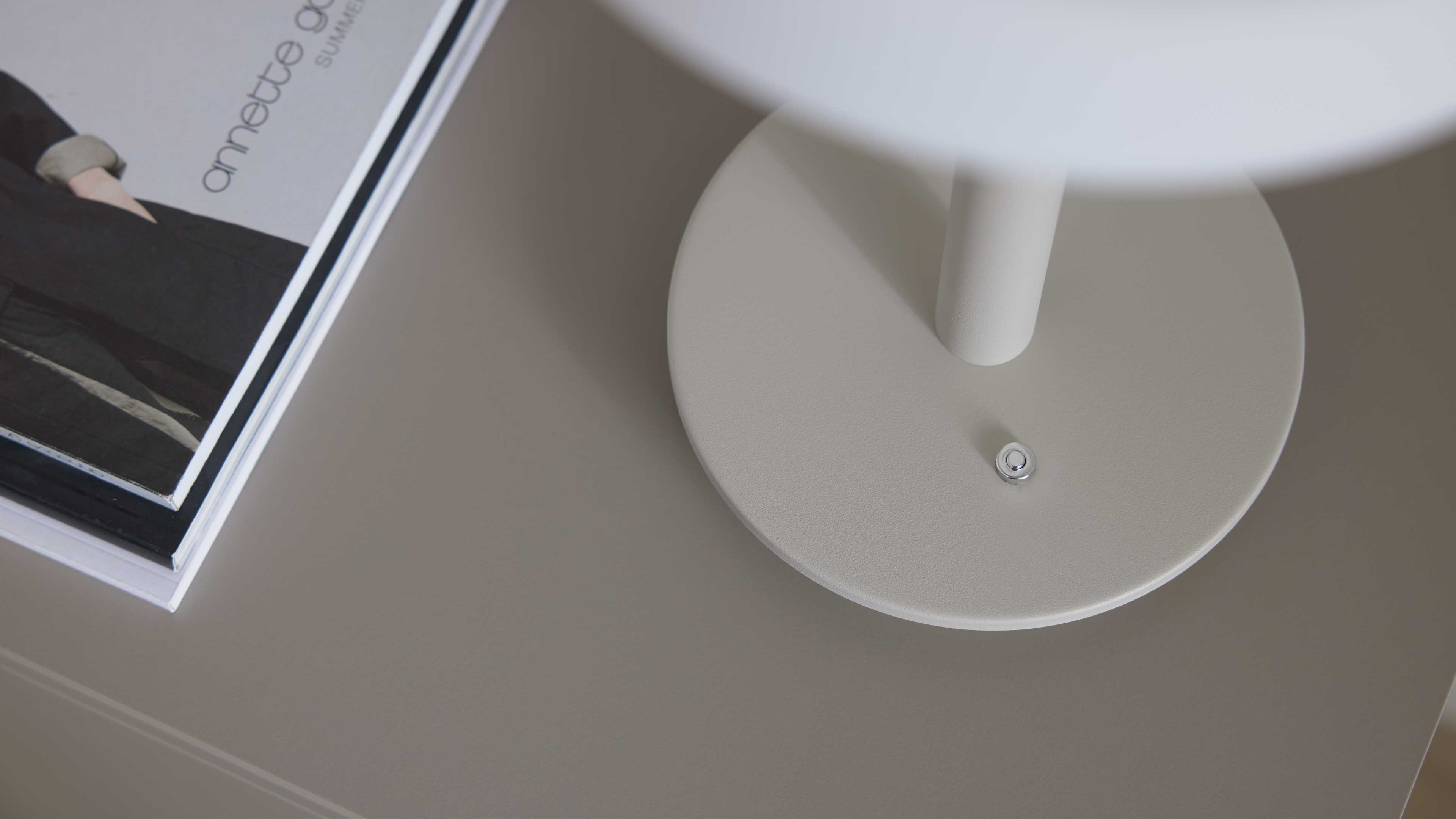 Runder, mattgrauer Lampensockel mit zylindrischem Stiel und kleinem runden Metallknopf auf grauem Tisch neben einem Stapel Zeitschriften.