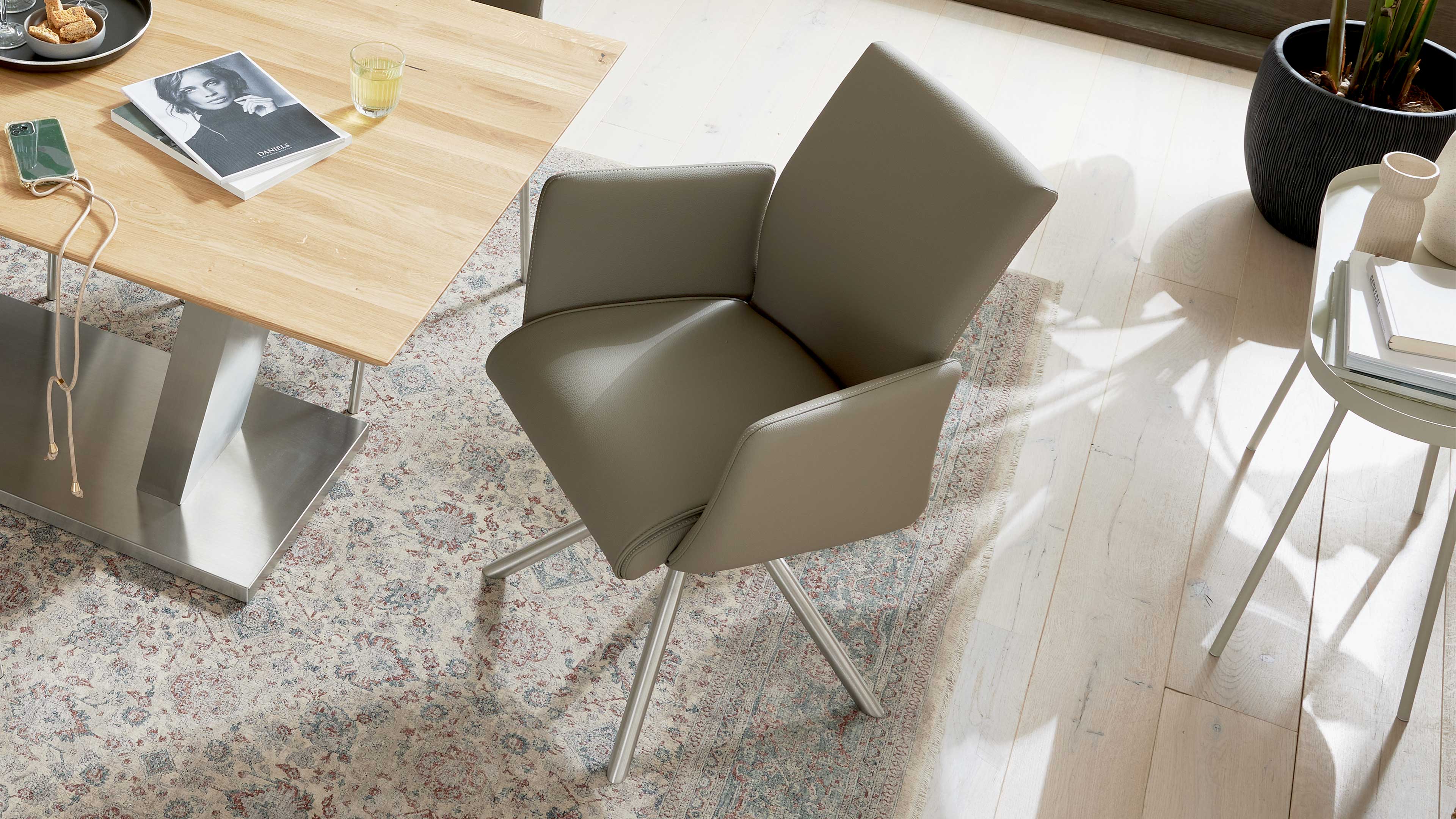 Gepolsterter taupefarbener Drehstuhl mit vier schlanken Metallbeinen auf gemustertem Teppich neben Holztisch.
