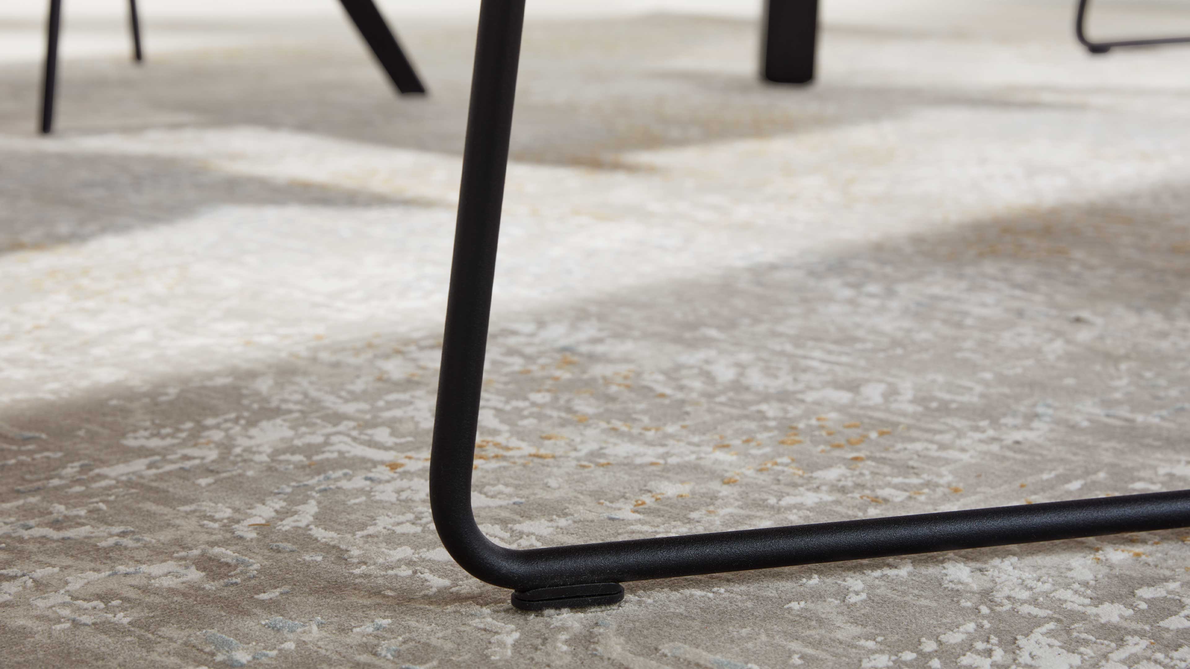 Schwarzes Rundrohr-Gestell eines Stuhl- oder Tischbeins auf hellgrau-beigem, strukturiert gemustertem Teppich.
