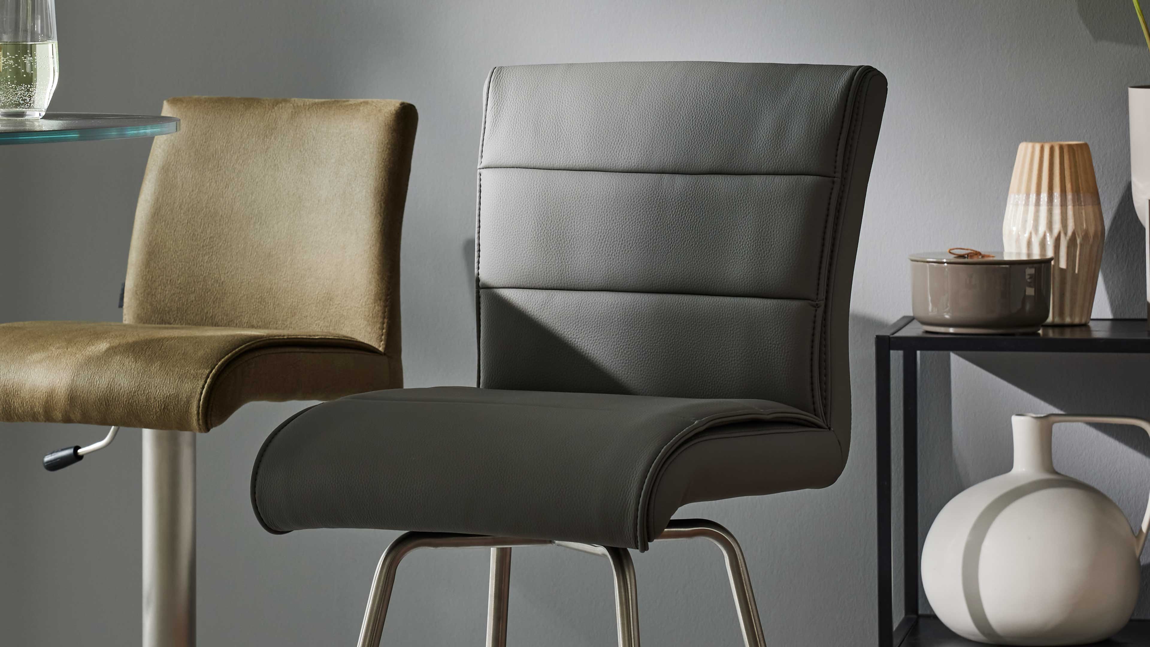 Dunkelgrauer, gepolsterter Barhocker mit Metallfuß im Vordergrund, links ein hellbrauner Stuhl, rechts ein Regal mit Keramikvasen.