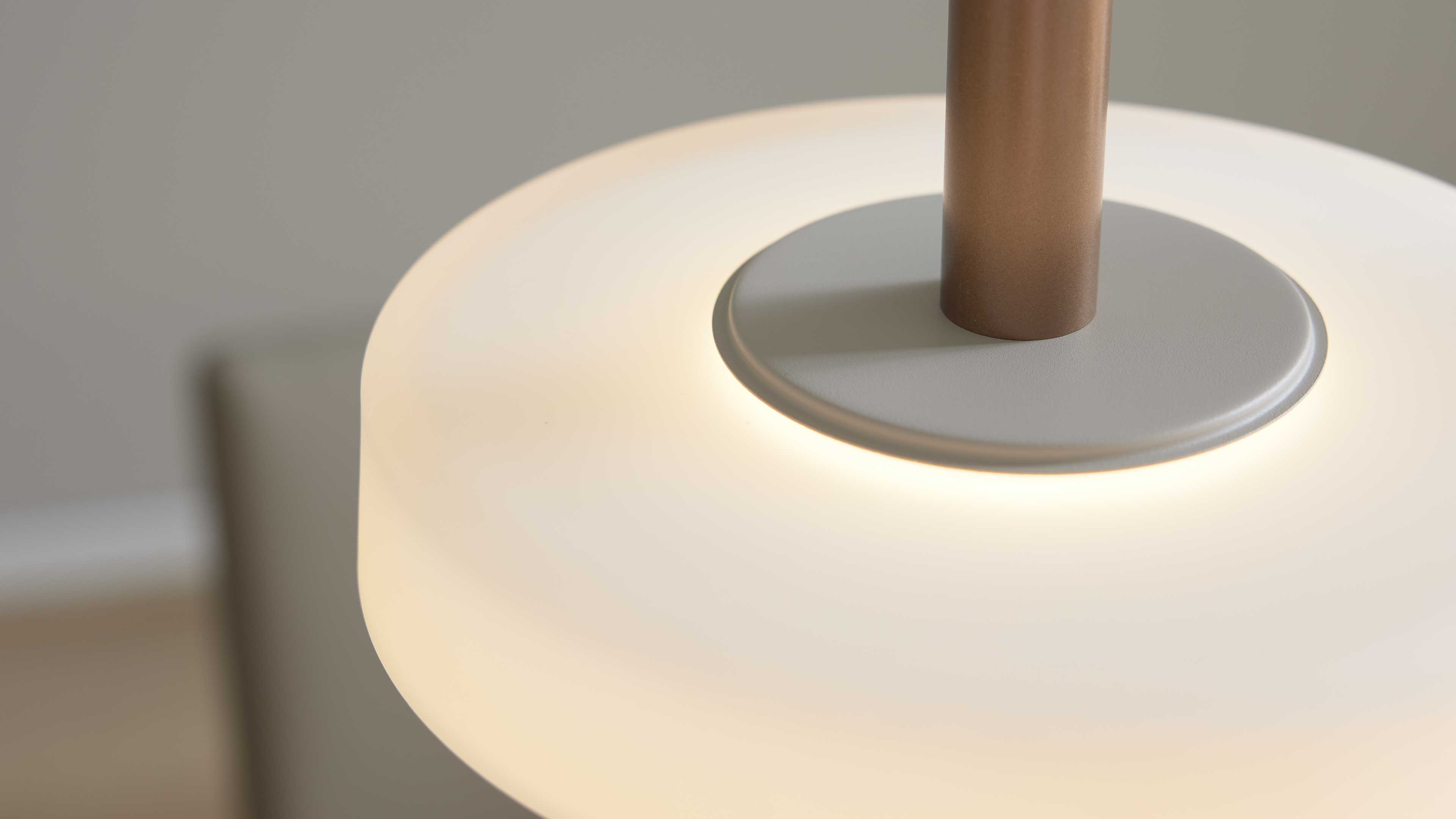 Nahaufnahme einer beleuchteten Pendelleuchte mit milchweißem Diffusor, grauer Abdeckung und matt kupferfarbener Stange.