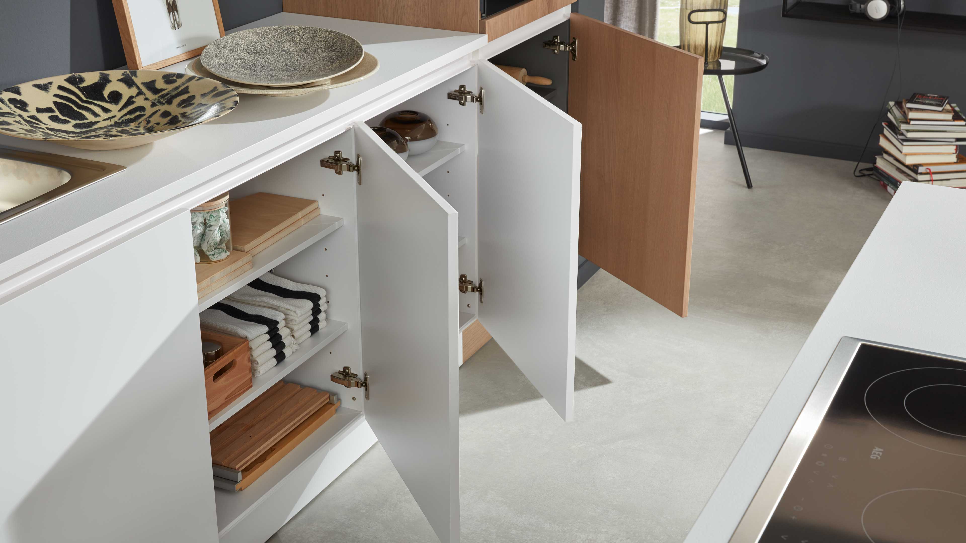 Geöffnete weiße Küchenunterschränke mit Holzfront, Innenregalen mit gefalteten Handtüchern und Holzbrettchen sowie Arbeitsplatte mit Spüle.