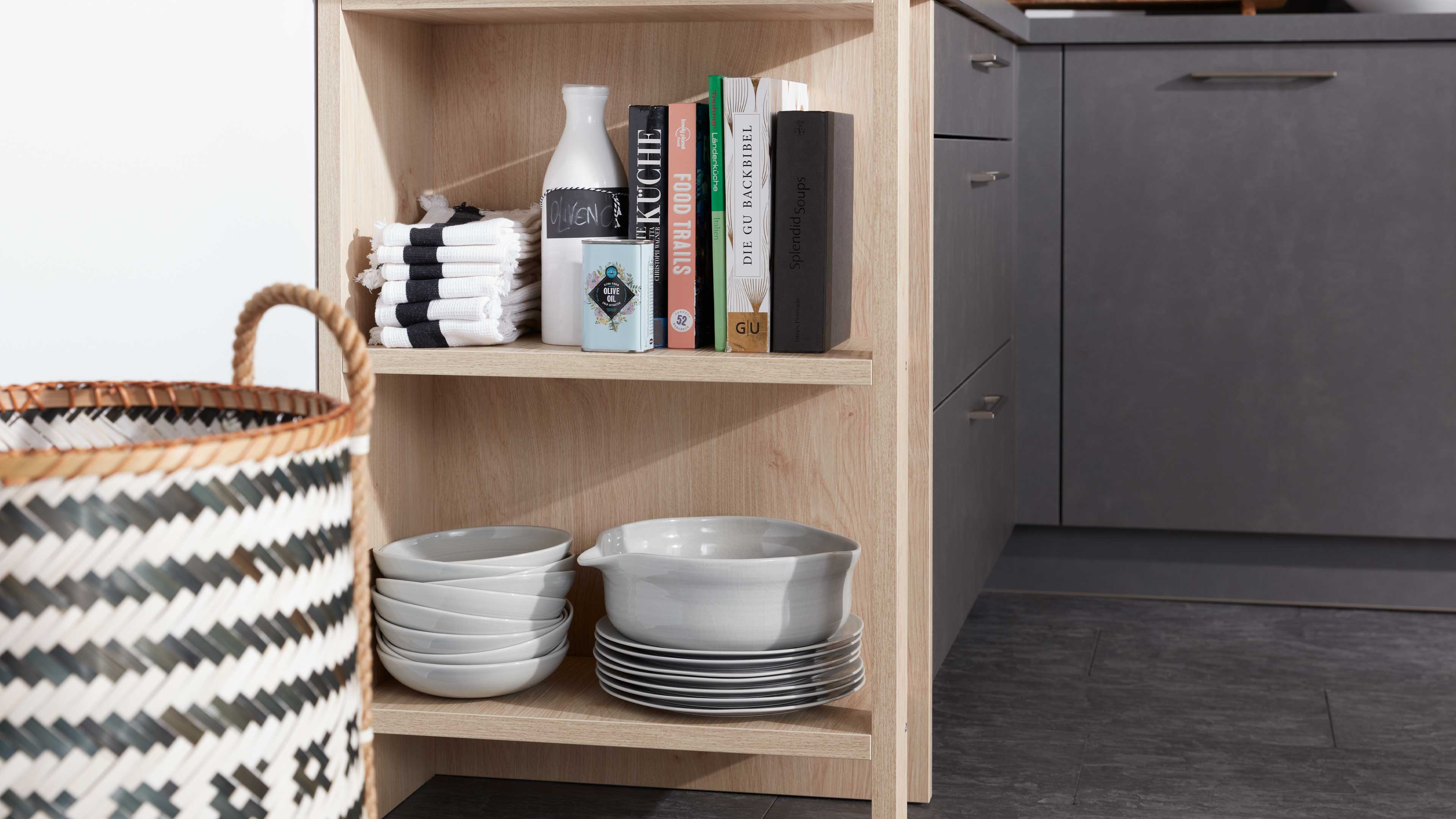 Offenes Regal in heller Holzoptik mit gestapelten weißen Keramikschalen und -tellern unten, oben Handtücher, Bücher und Dosen.
