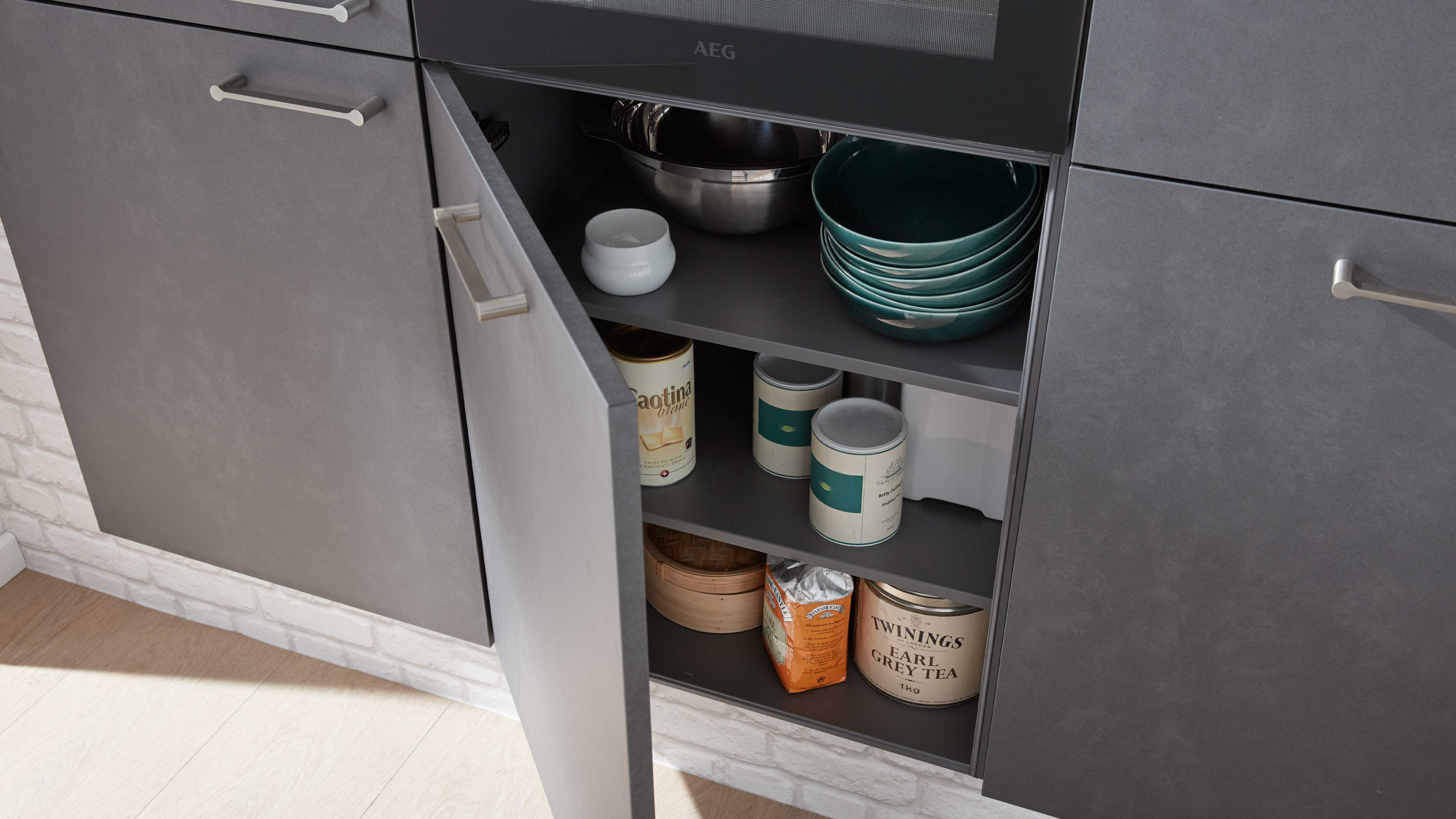 Geöffneter grauer Küchenunterschrank mit offenem Fach, darin Stapel türkiser Schalen, Edelstahlschüssel, Dosen und ein rundes Holzgefäß.