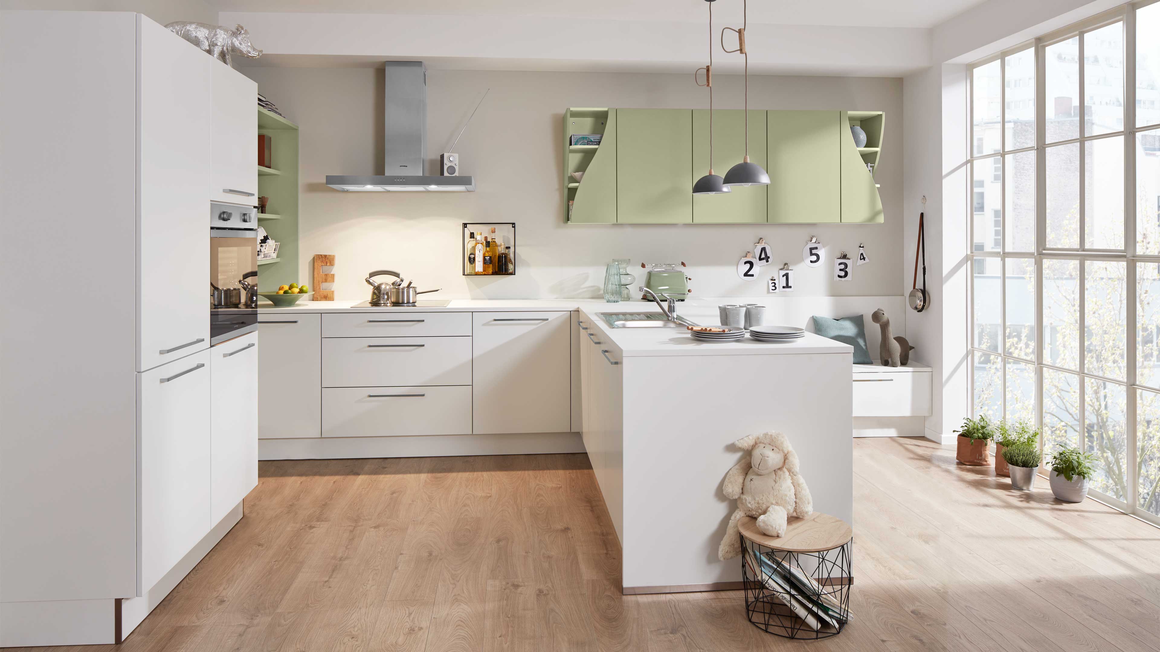 Offene, helle Küche mit weißen Unterschränken, pastellgrünen Hängeschränken, Kochinsel mit Spüle, Holzboden und großen Fenstern.