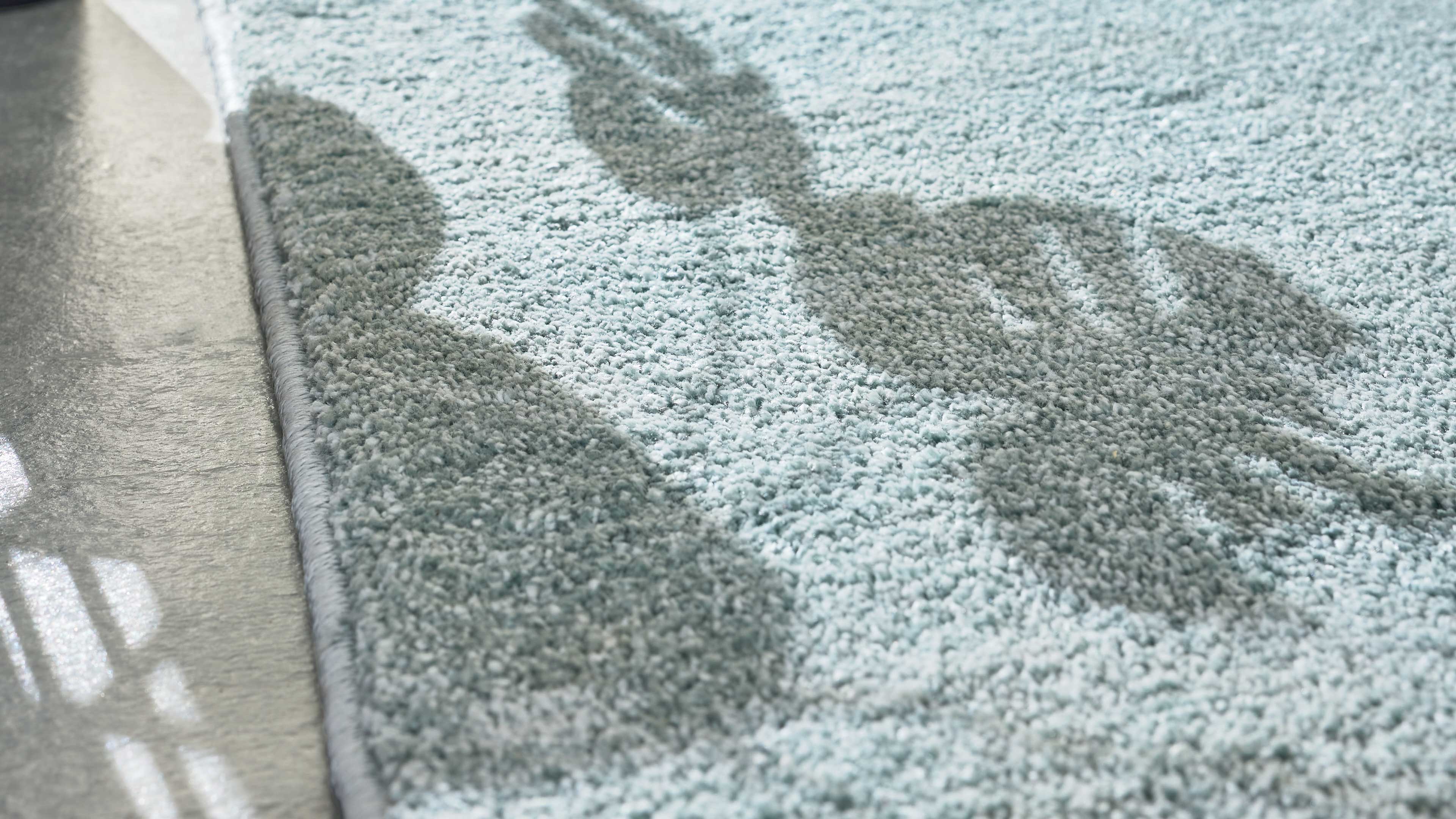 Nahaufnahme eines hellblauen, flauschigen Teppichs mit sichtbarer Umkettelung, blattförmigen Schatten und angrenzendem Bodenfliesen.