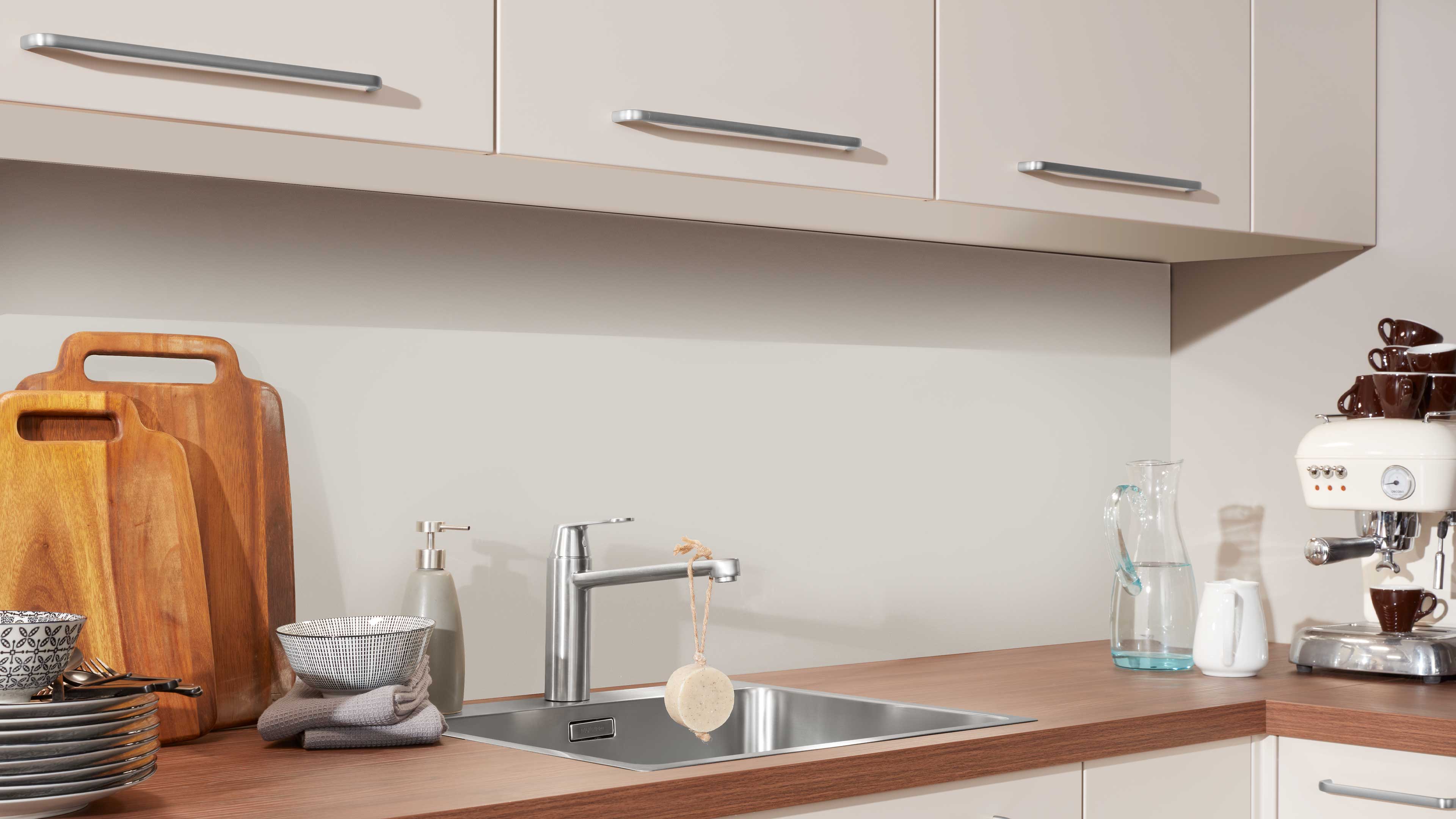 Eingelassener Edelstahl-Spülbecken mit schlankem Chrom-Einhebelmischer auf Holzarbeitsplatte, daneben Schneidebretter und Geschirr.