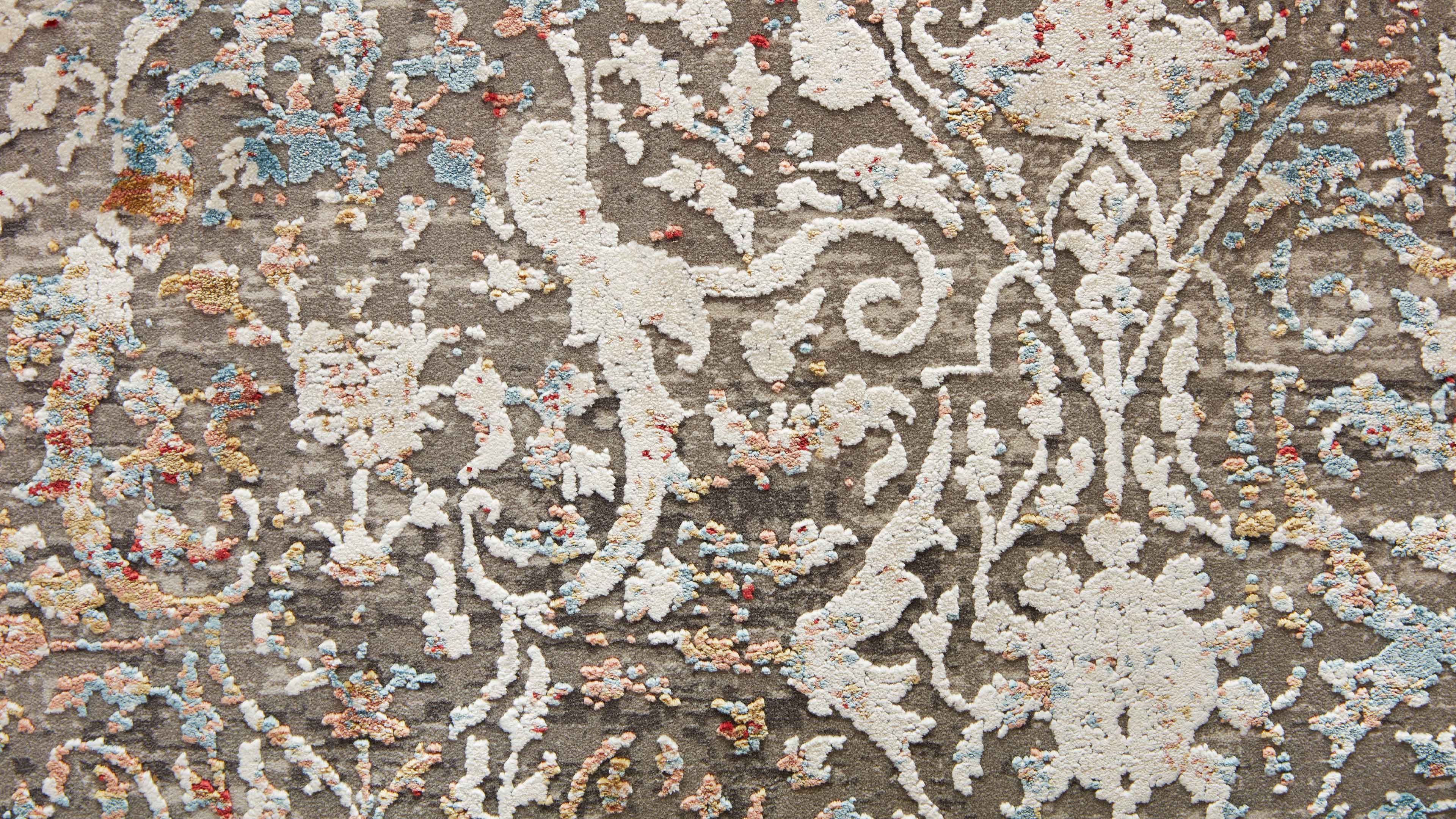 Teppich in sandbeige mit reliefartigem weißen Ranken- und Blumenmuster sowie verstreuten pastellfarbenen Garnknoten.