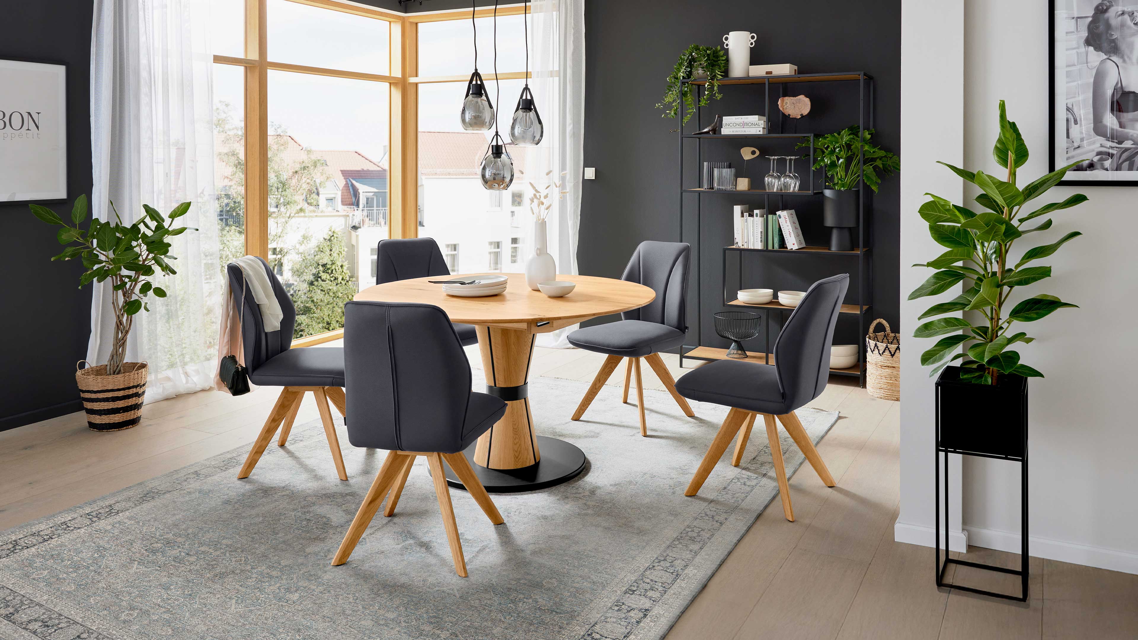 Runder Holz-Esstisch mit fünf grauen, gepolsterten Stühlen auf Holzbeinen, daneben Regal, Hängeleuchten und Zimmerpflanzen.
