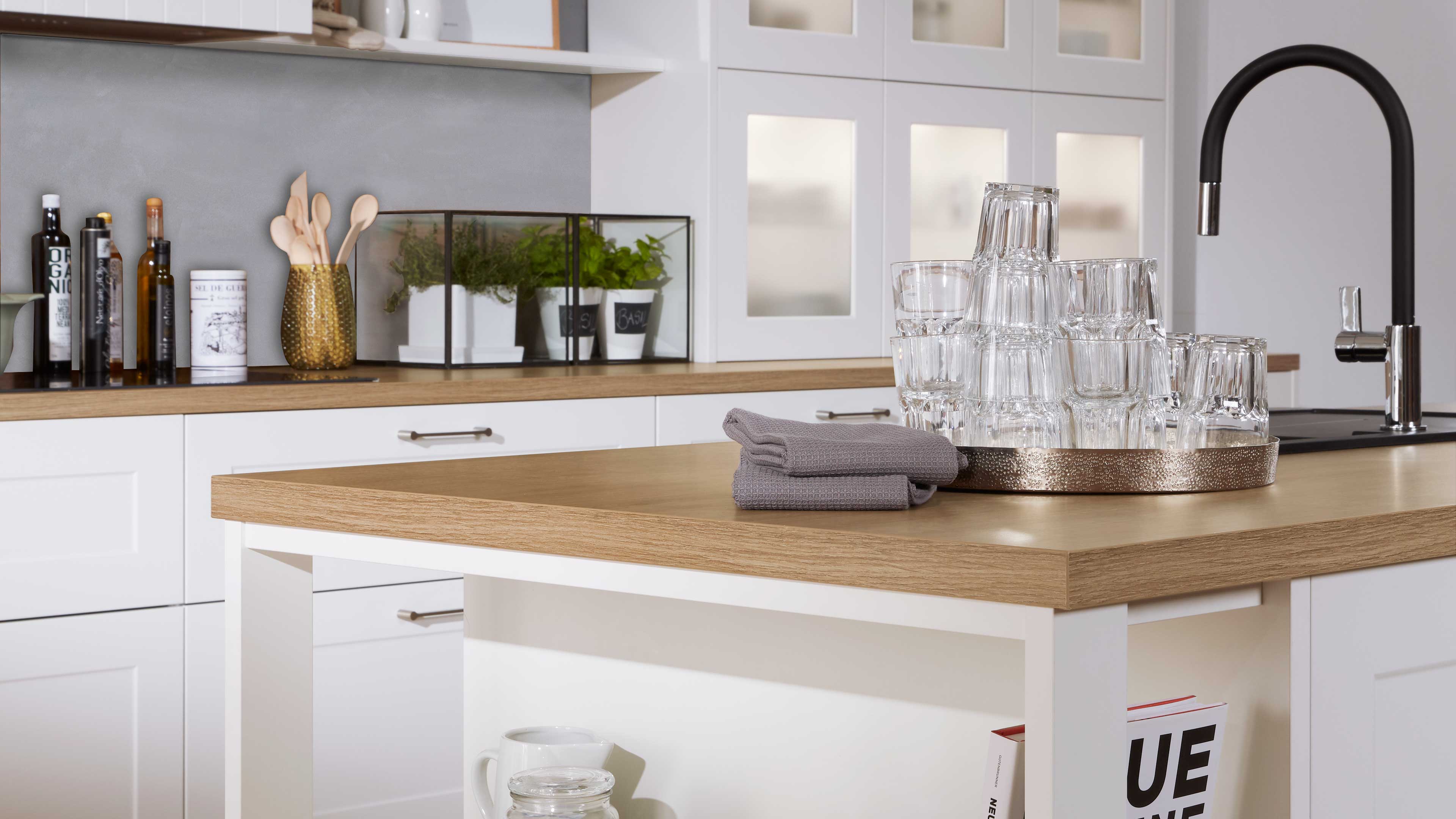 Kücheninsel mit heller Holzarbeitsplatte und weißem Korpus, darauf gefaltete graue Geschirrtücher und ein Tablett mit gestapelten Gläsern.